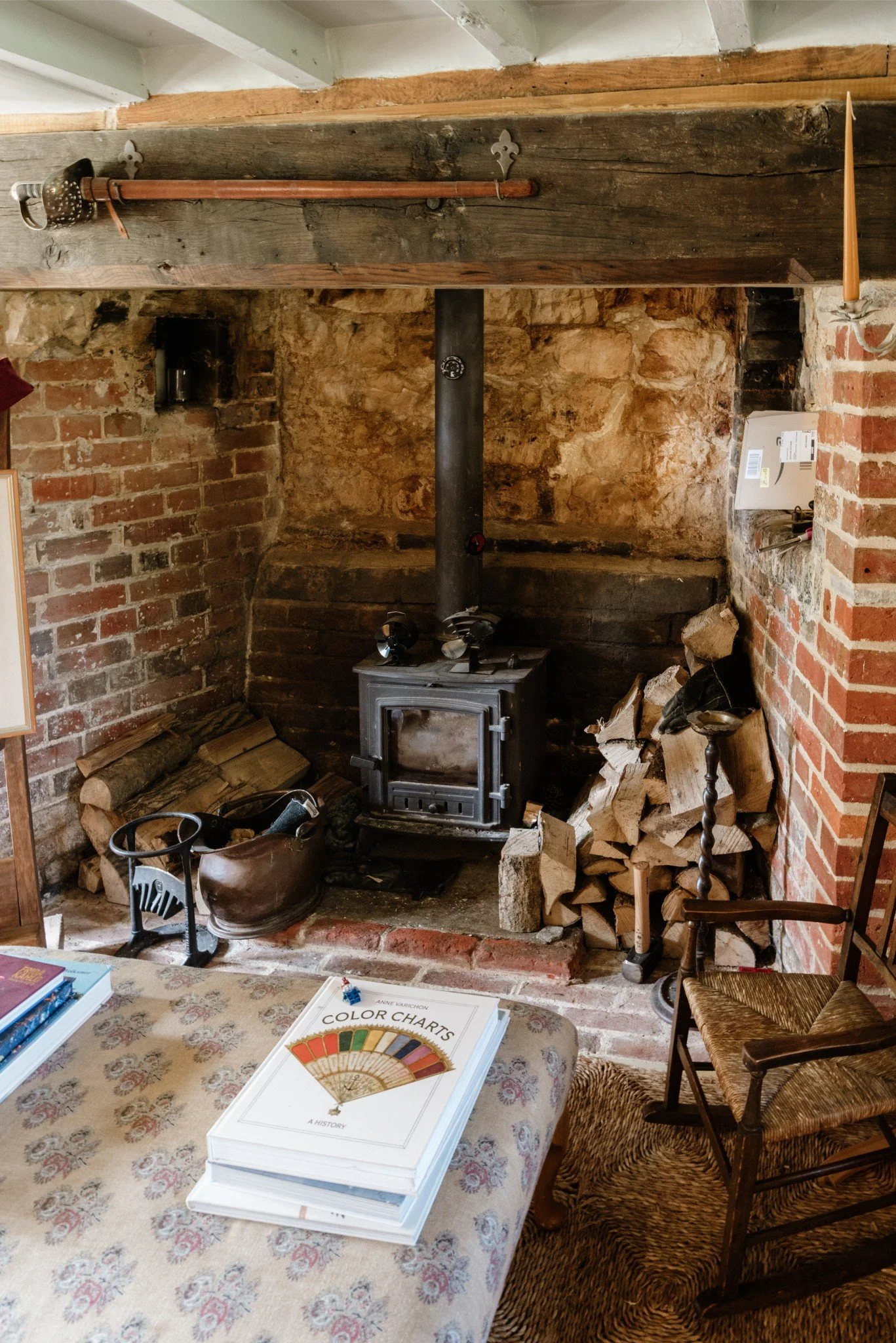 A cozy room featuring a vintage wood stove with a pipe extending upward, surrounded by stacked firewood. There is a table in the foreground with books and papers, including a book titled 'Color Charts'. A wicker and wood chair is positioned near the 