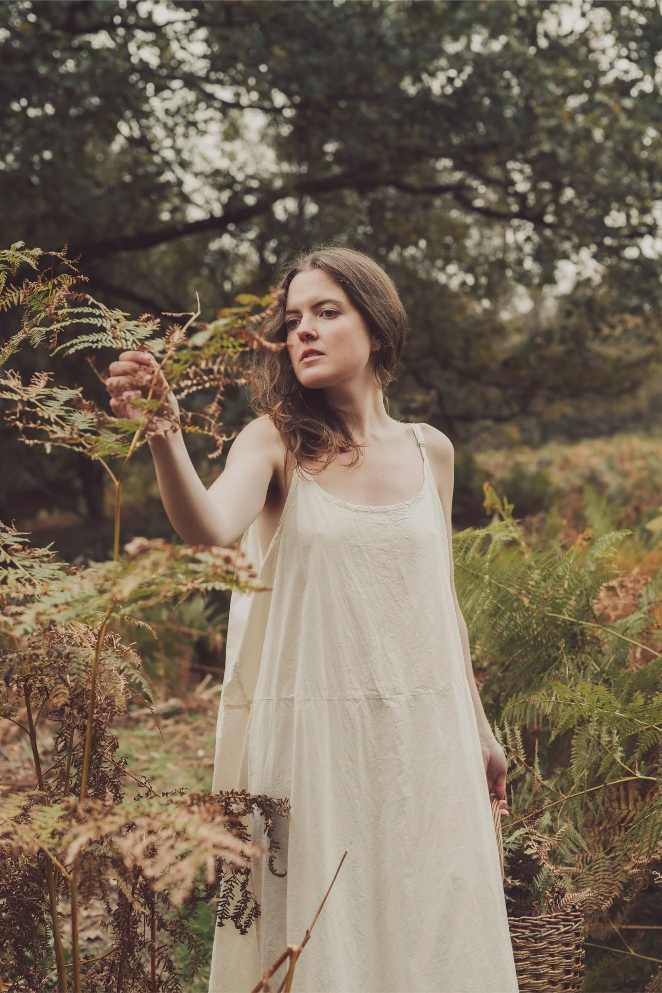 A woman in a cream-colored dress stands outdoors among ferns and trees, gently touching a fern with her hand, with a contemplative expression.