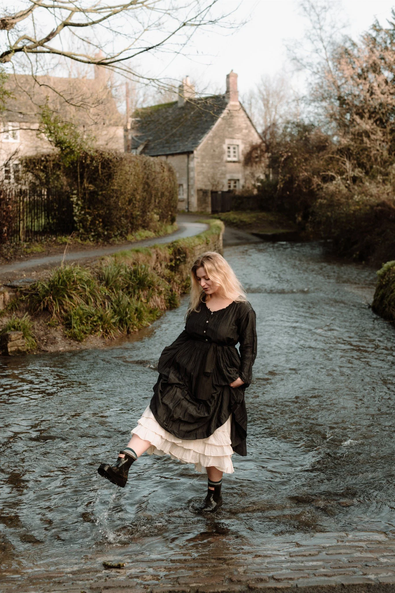 Woman standing in a river, wearing a black dress, cream-colored ruffled skirt, and black boots, with a rural house and trees in the background.