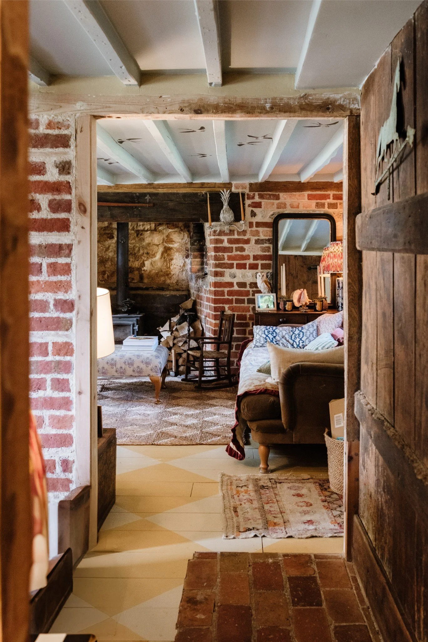 View through a rustic doorway into a cozy living room with brick walls, a wood stove, a fireplace with firewood, a rocking chair, a sofa with multiple pillows, a table with a lamp, and a mirror on the wall.