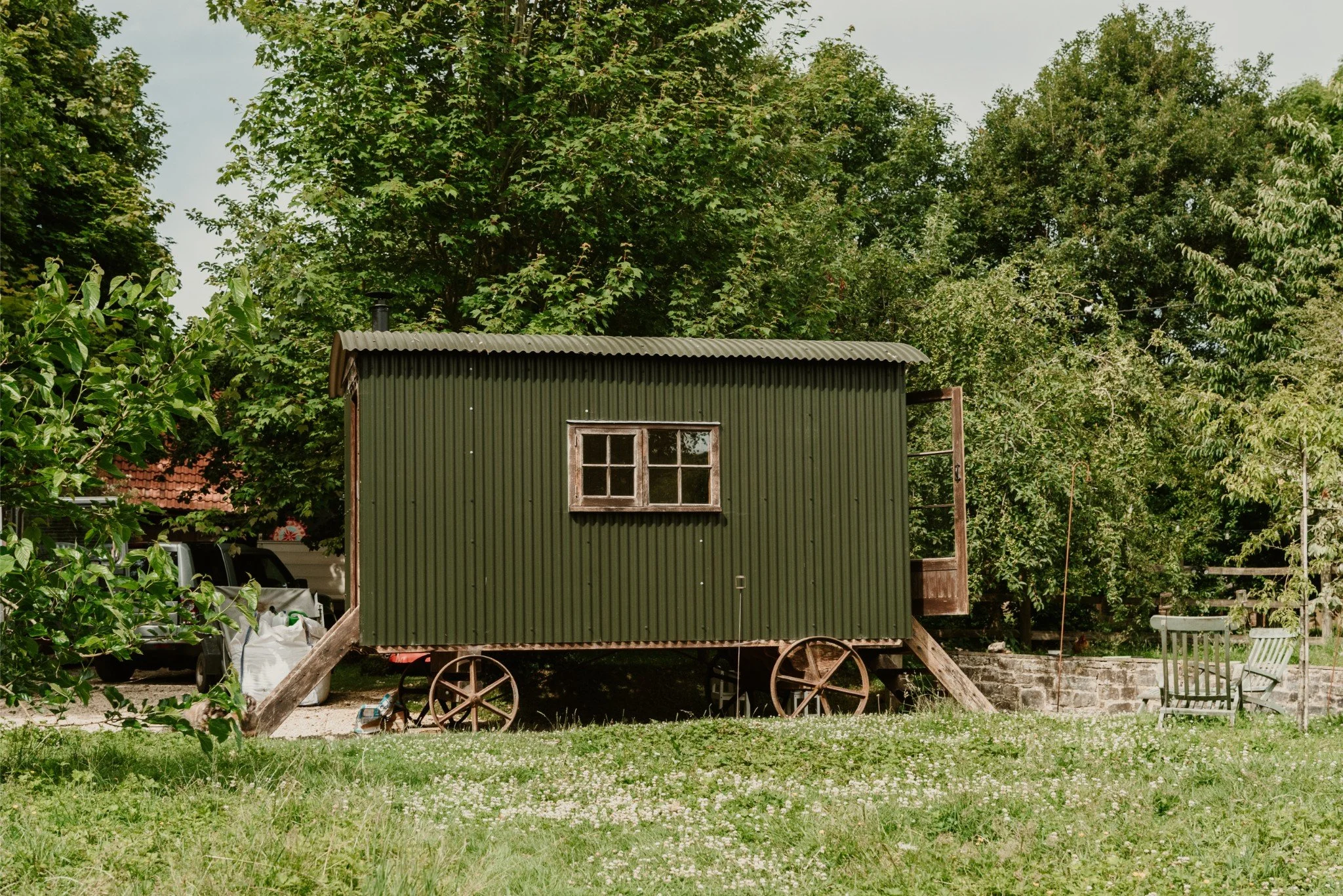 A small, green, corrugated metal tiny house on wheels with a window, set on a grassy area with trees and chairs nearby.