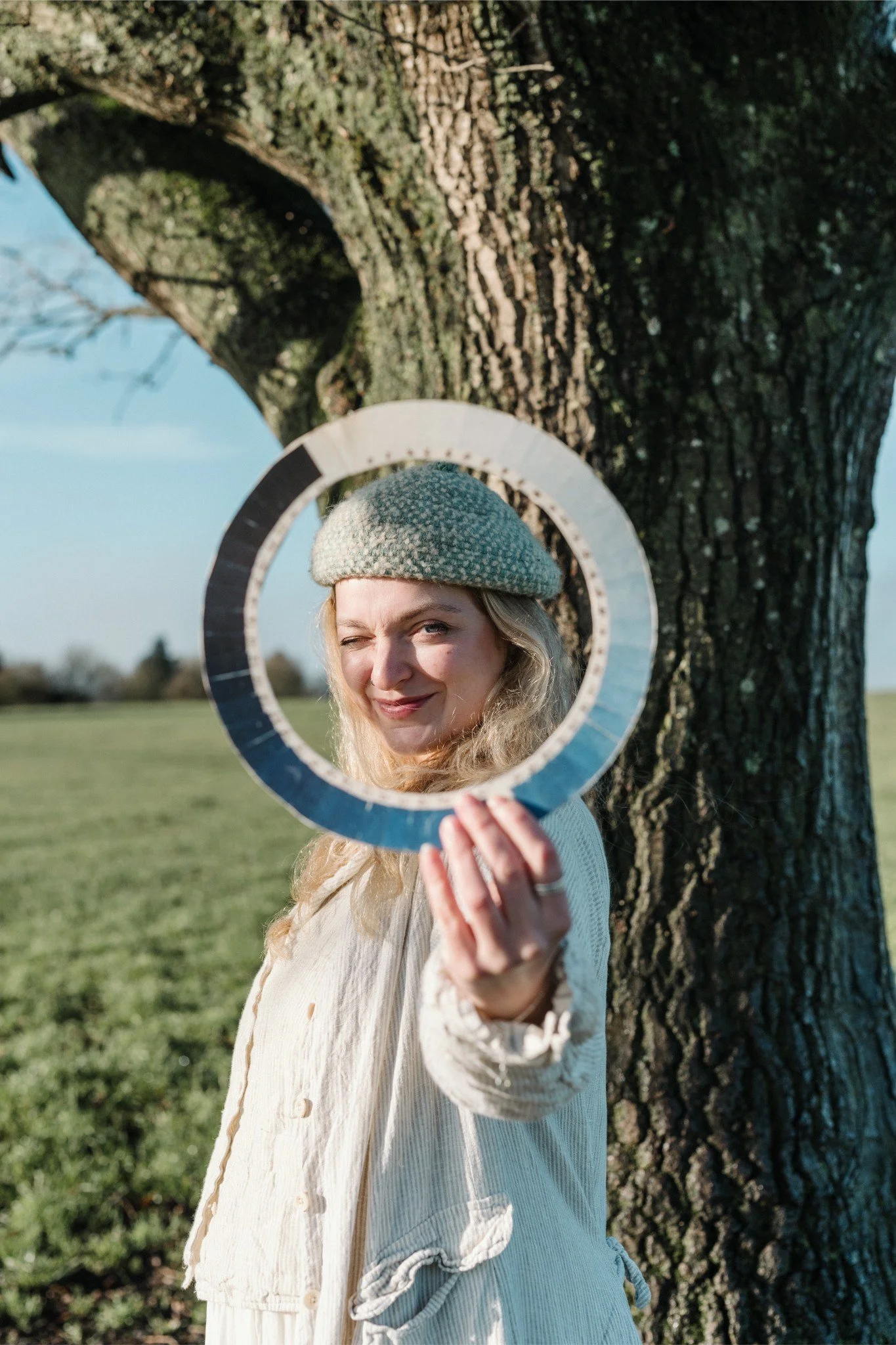 Woman standing outdoors by a large tree, holding a circular reflective object in front of her face, winking and smiling, wearing a knitted beret and a light-colored jacket.