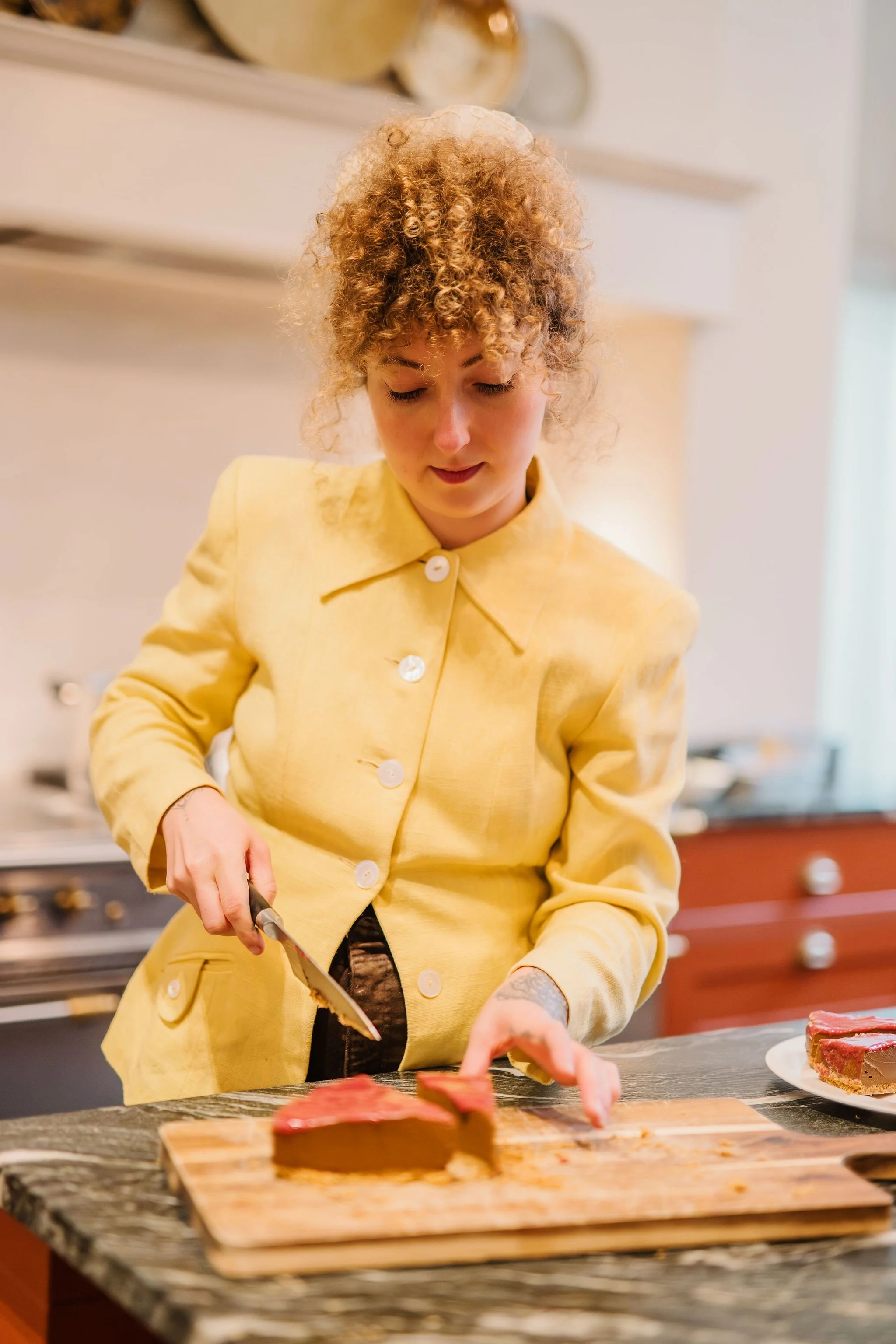 A woman with curly hair, wearing a yellow jacket, is slicing a piece of meat on a wooden cutting board in a kitchen.