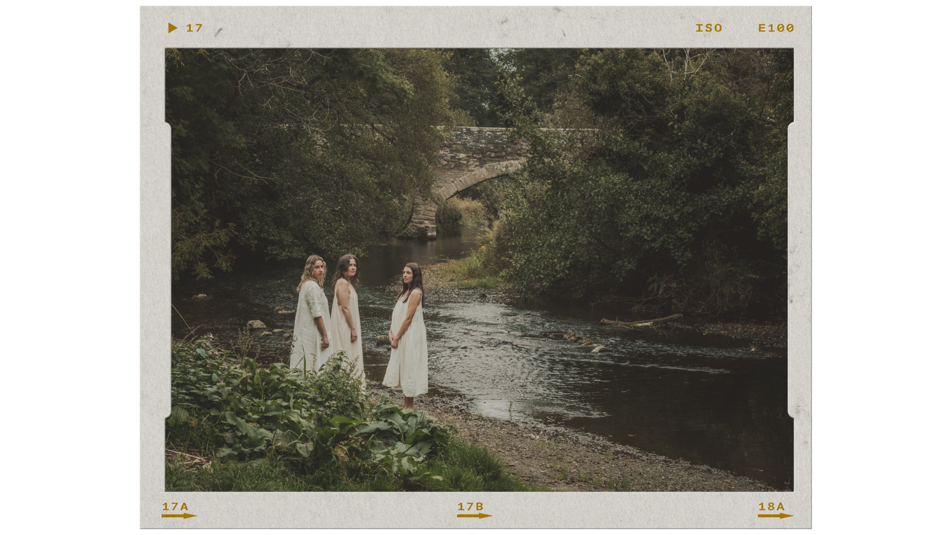 Three women wearing white dresses standing by a river with trees and an old stone bridge in the background.