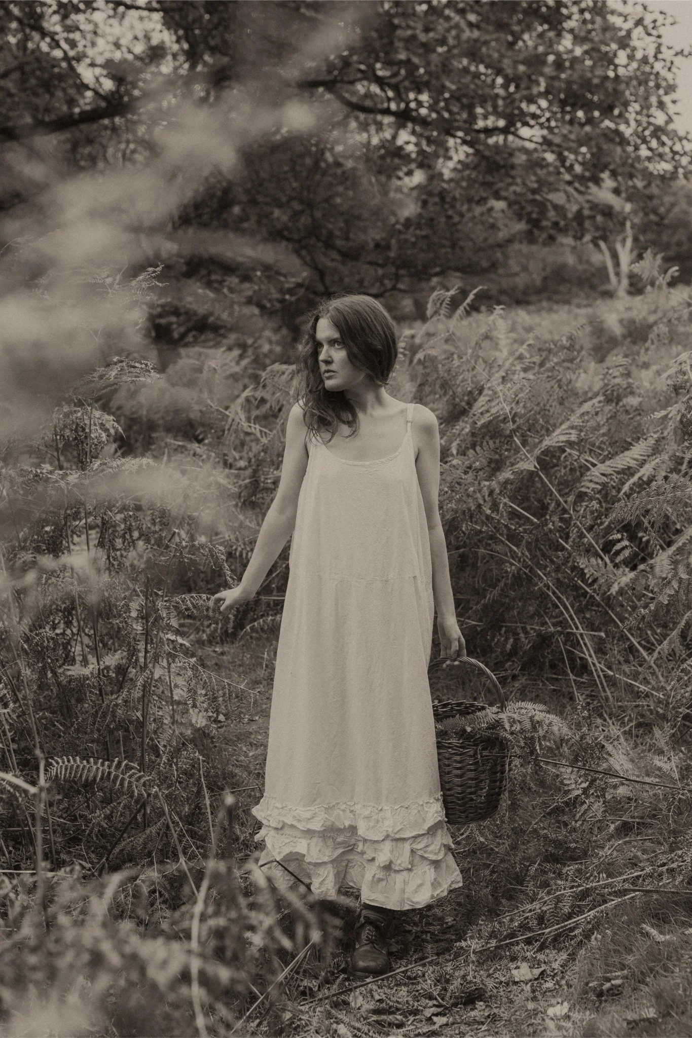 A young woman in a long, flowing dress walking through a wooded area, holding a basket, in black and white.