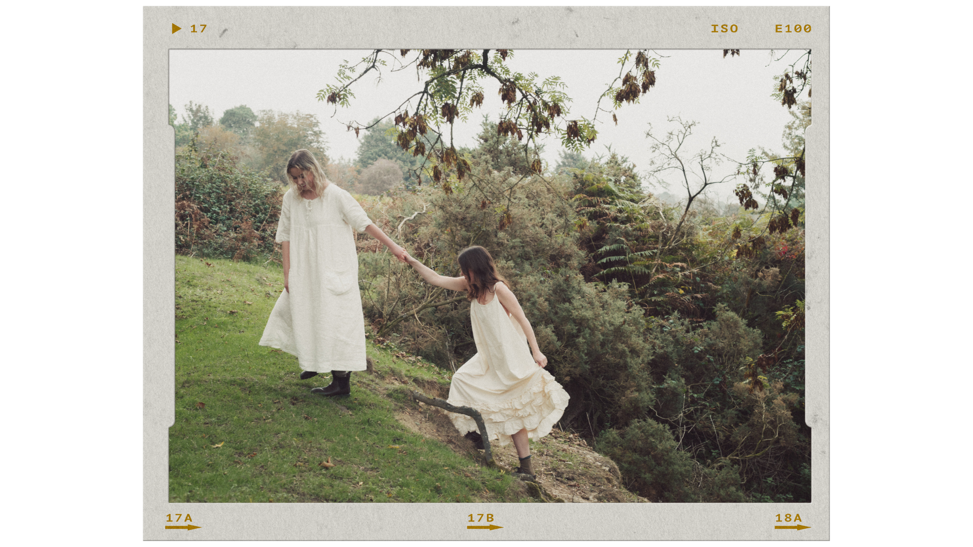 Two girls in white dresses, one helping the other as she steps down a grassy incline in a natural outdoor setting with trees and shrubs.