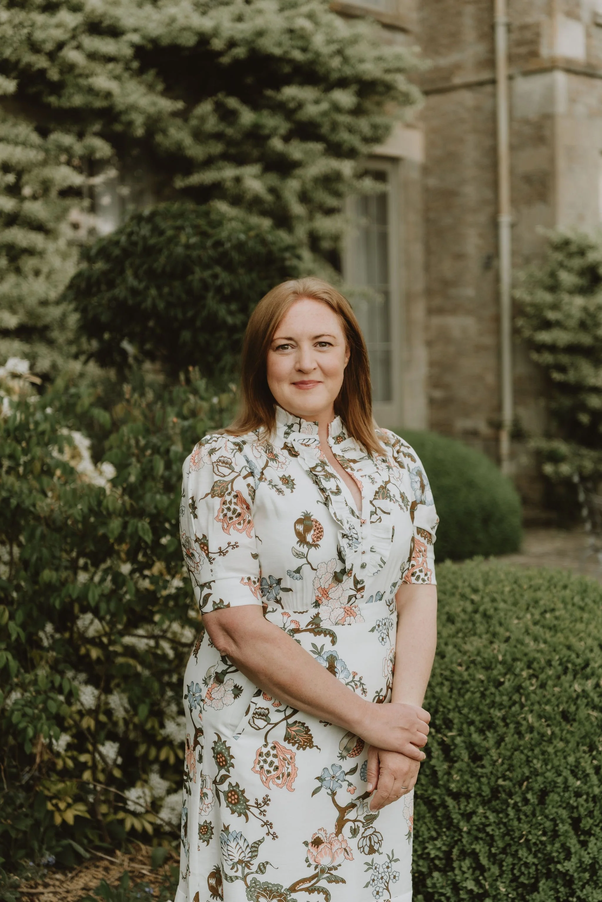 A woman with shoulder-length reddish-brown hair, wearing a white dress with a colorful floral pattern, standing outdoors in front of a garden with shrubbery and a building in the background.