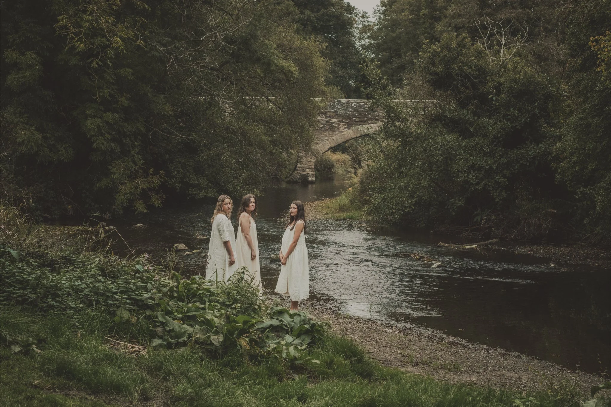 Three women in white dresses standing on the bank of a river surrounded by trees, with a stone bridge in the background.