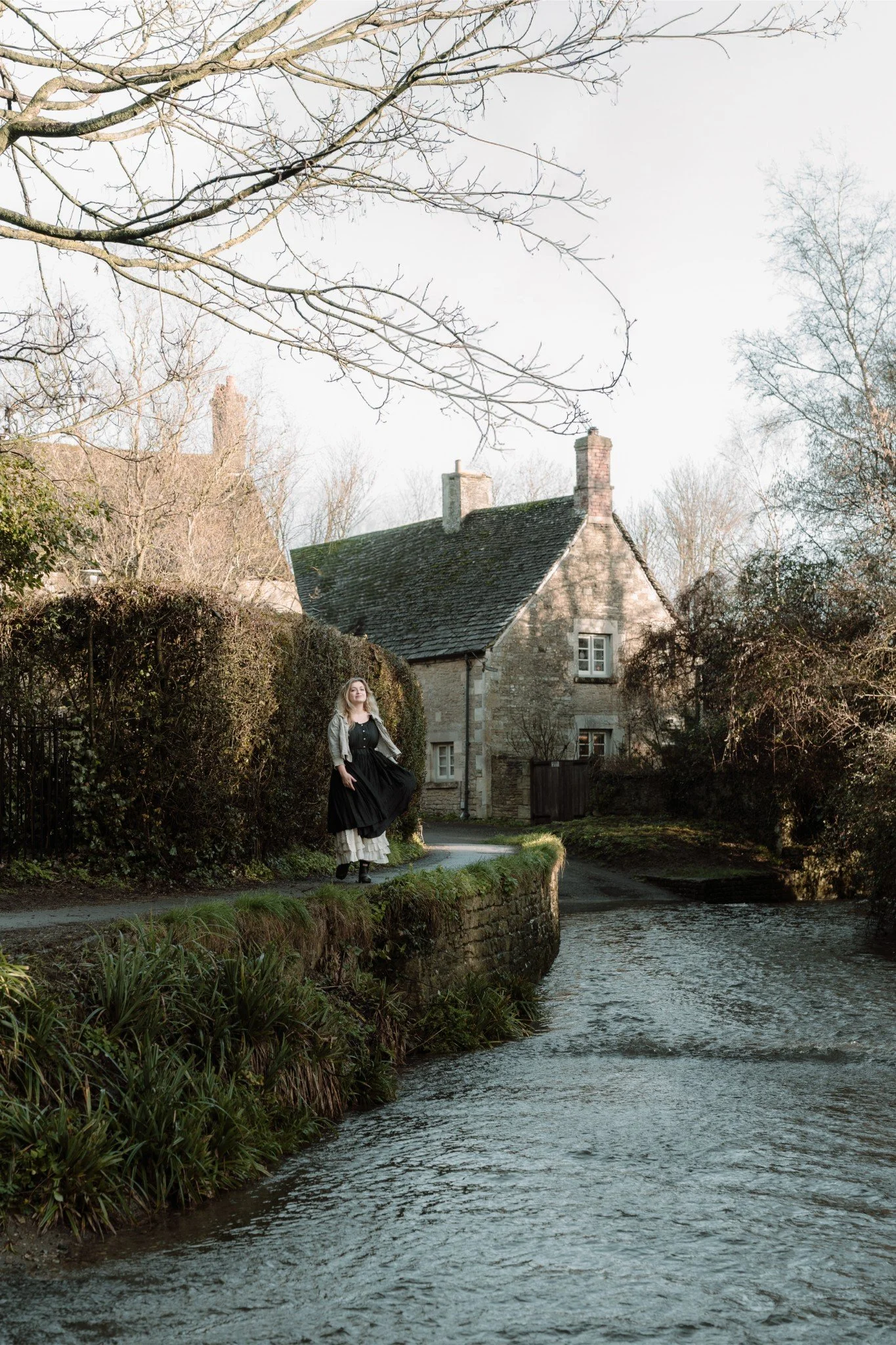 A woman standing on a narrow stone path next to a small river, with a stone house in the background and trees around, during daylight.