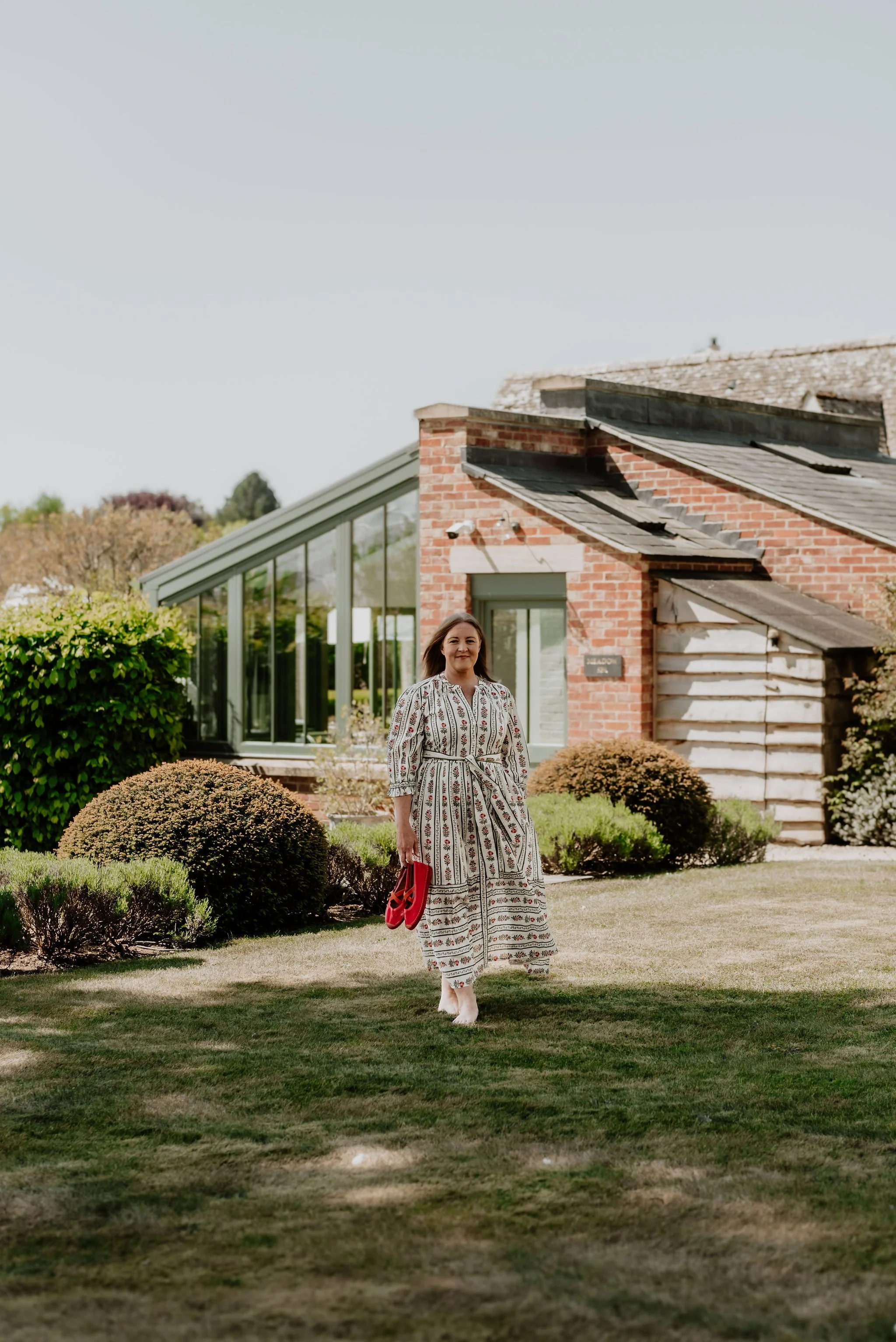 A woman in a floral dress holding a red purse standing on a well-kept lawn in front of a modern glass and brick building with shrubs.