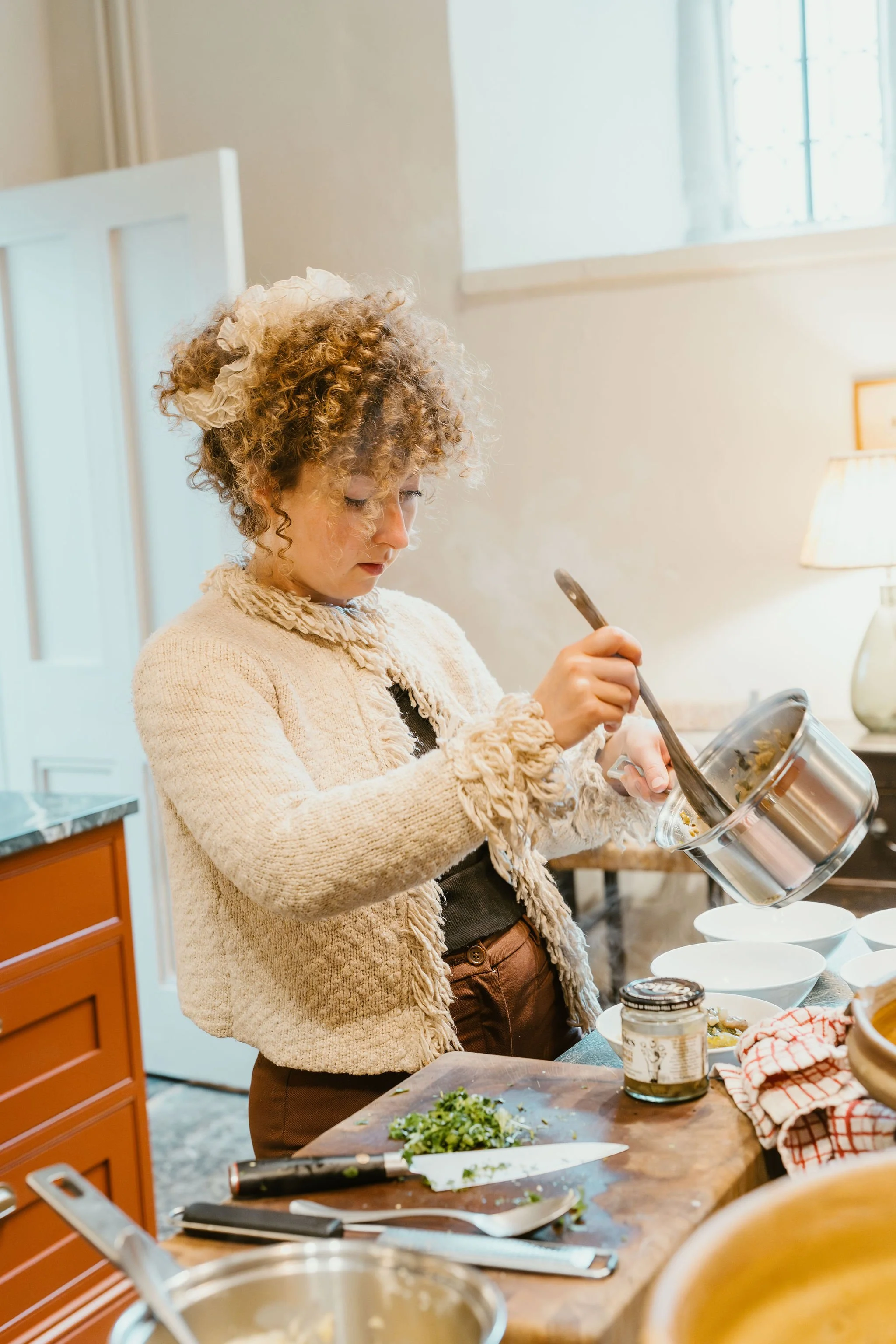 Woman chef with curly hair and a beige sweater preparing food in a kitchen.