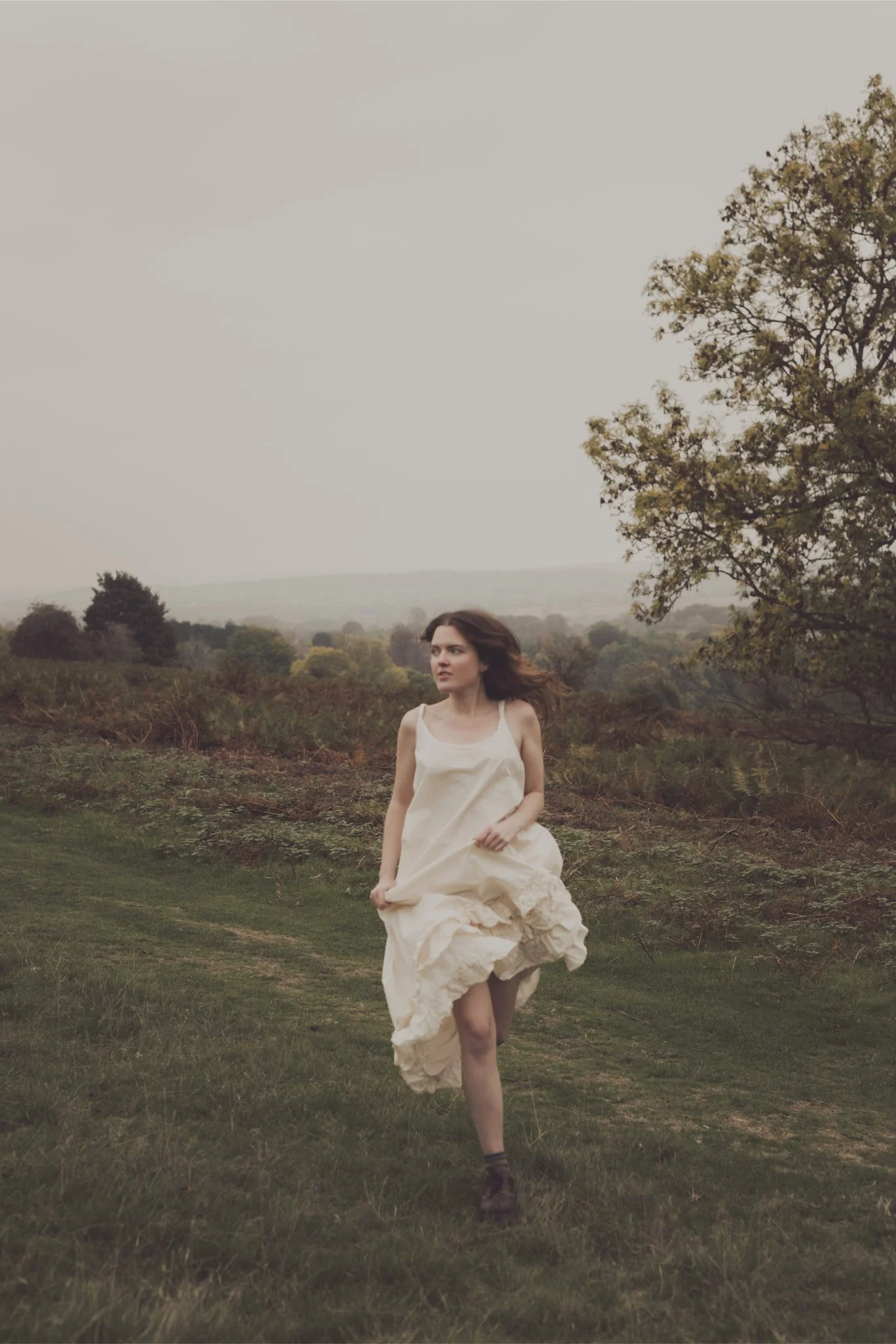 A woman running outdoors on a grassy field with trees and hills in the background, under an overcast sky.