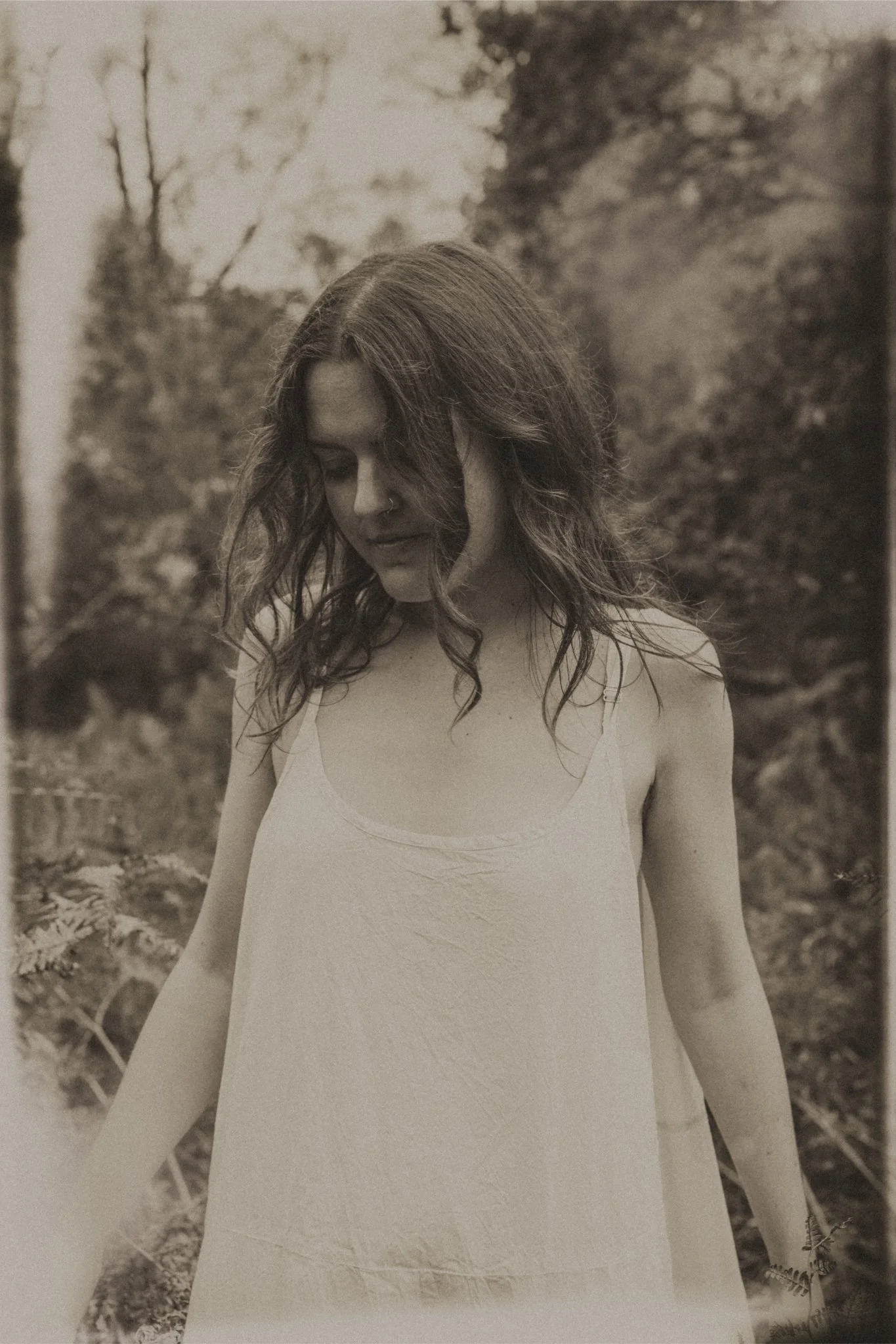 A woman with wavy hair wearing a loose, sleeveless dress standing outdoors in a natural setting with trees and foliage.