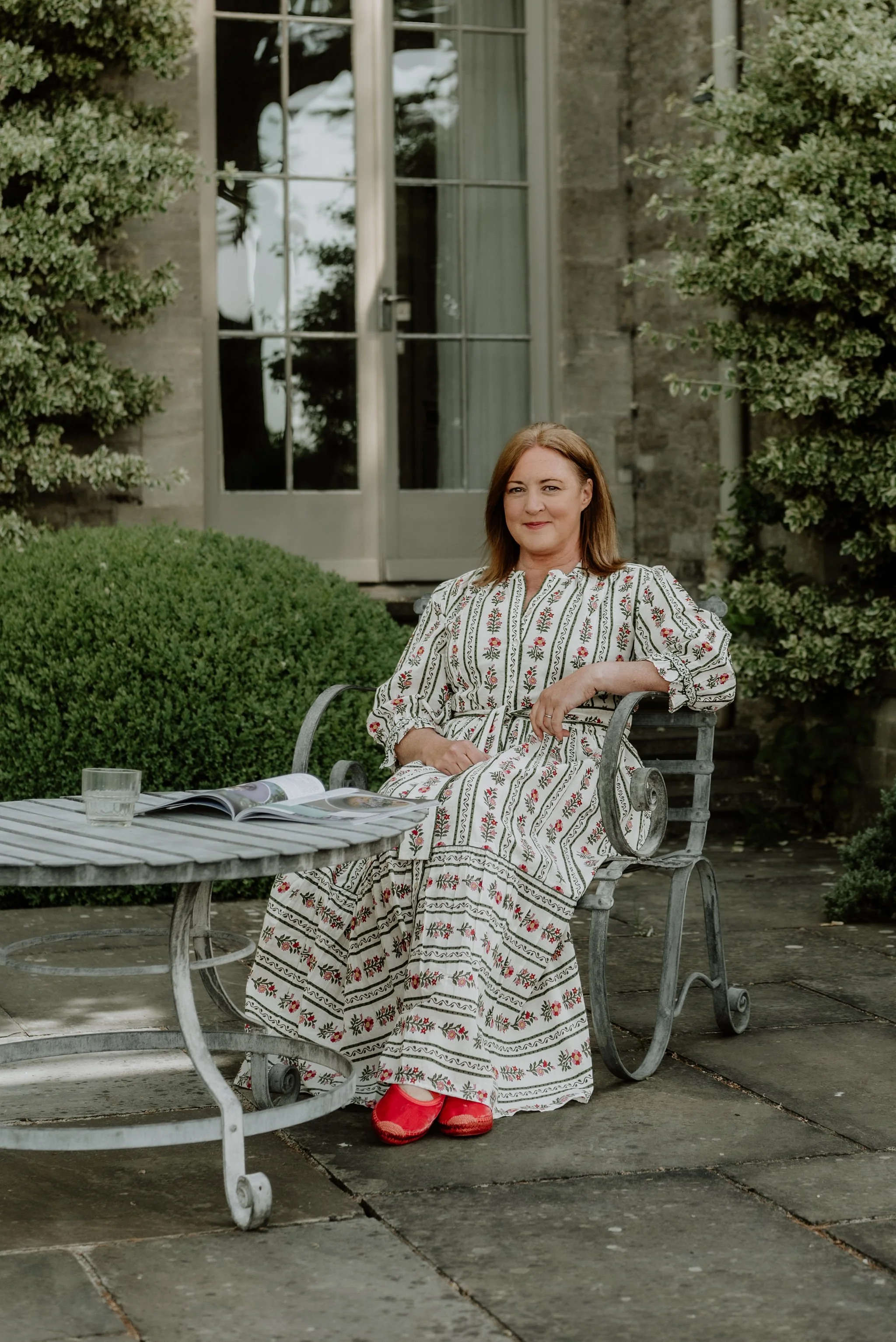 A woman in a long, white dress with embroidered floral patterns, sitting on a metal garden bench outdoors, with a glass of water and an open magazine on a nearby table, surrounded by greenery and a building with windows in the background.