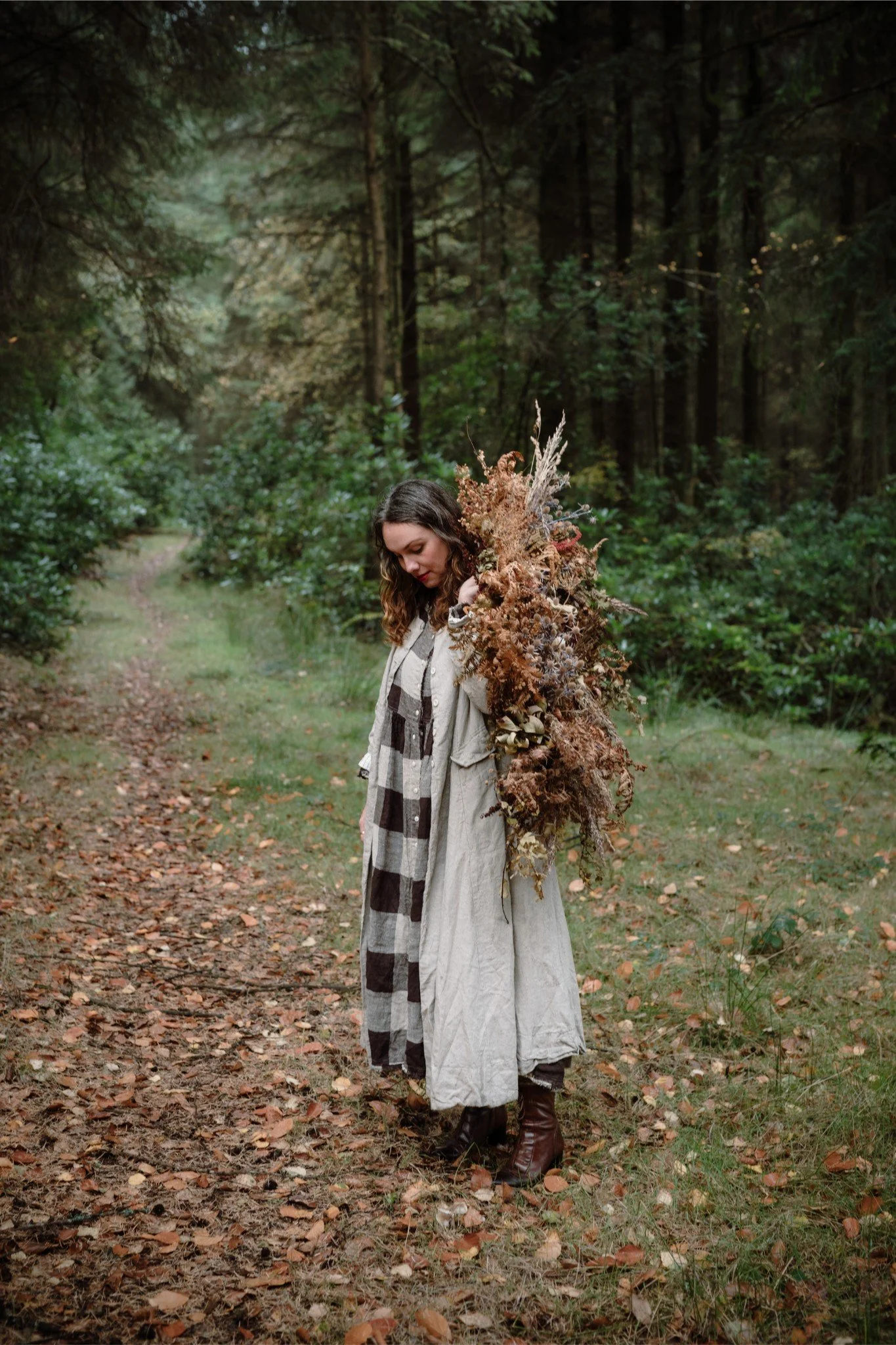 A woman standing on a forest path, holding a large bouquet of dried flowers and plants, wearing a long beige coat, brown boots, and a checkered scarf.