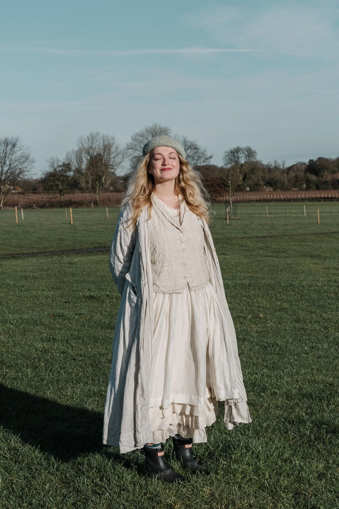 A young woman with long blonde hair standing outdoors in a green field, with her eyes closed and a content smile on her face, dressed in vintage-style beige and cream clothing, wearing a beret and rubber boots, under a blue sky.