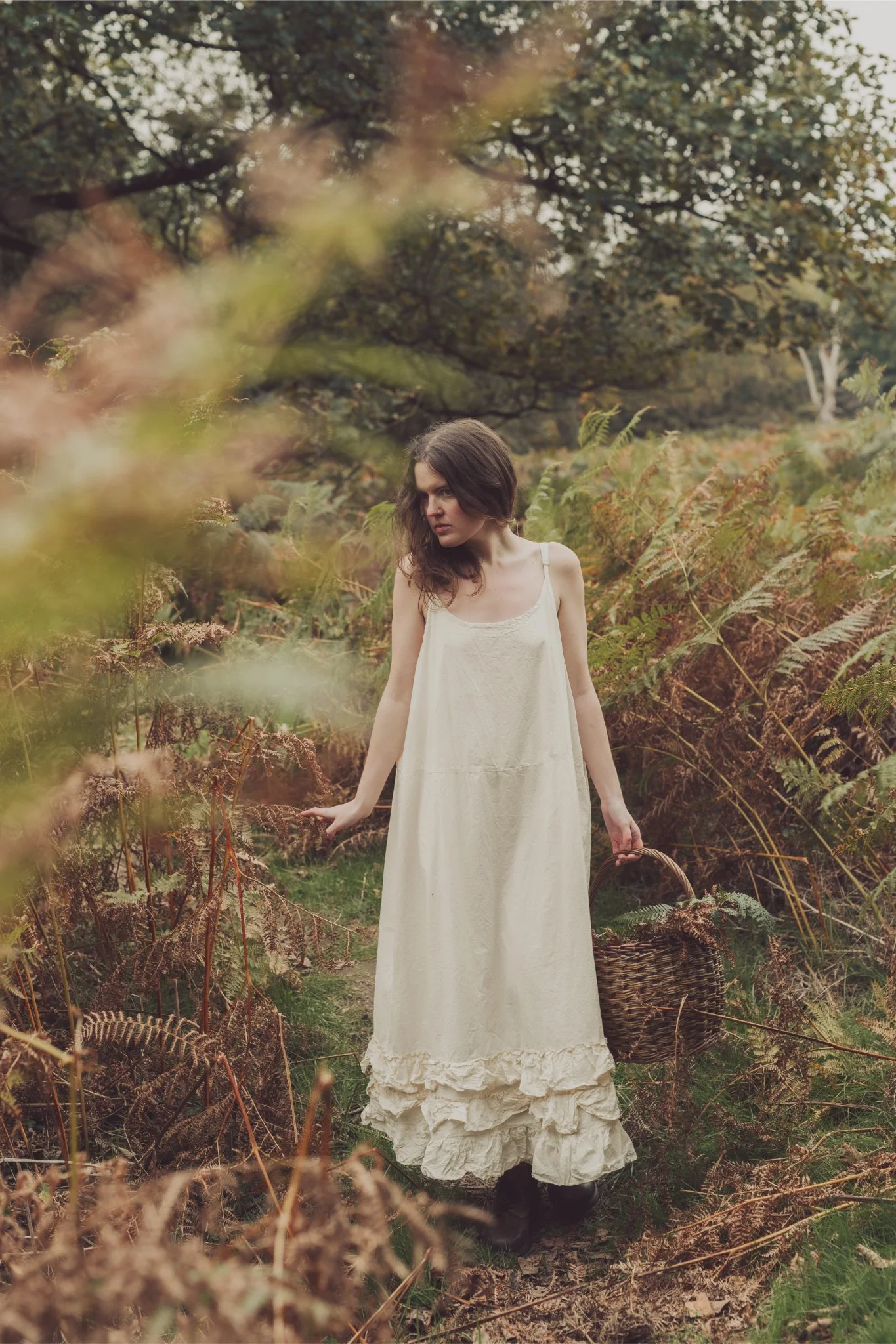 A young woman in a long, cream-colored dress standing in a forested area with ferns and trees, holding a basket.