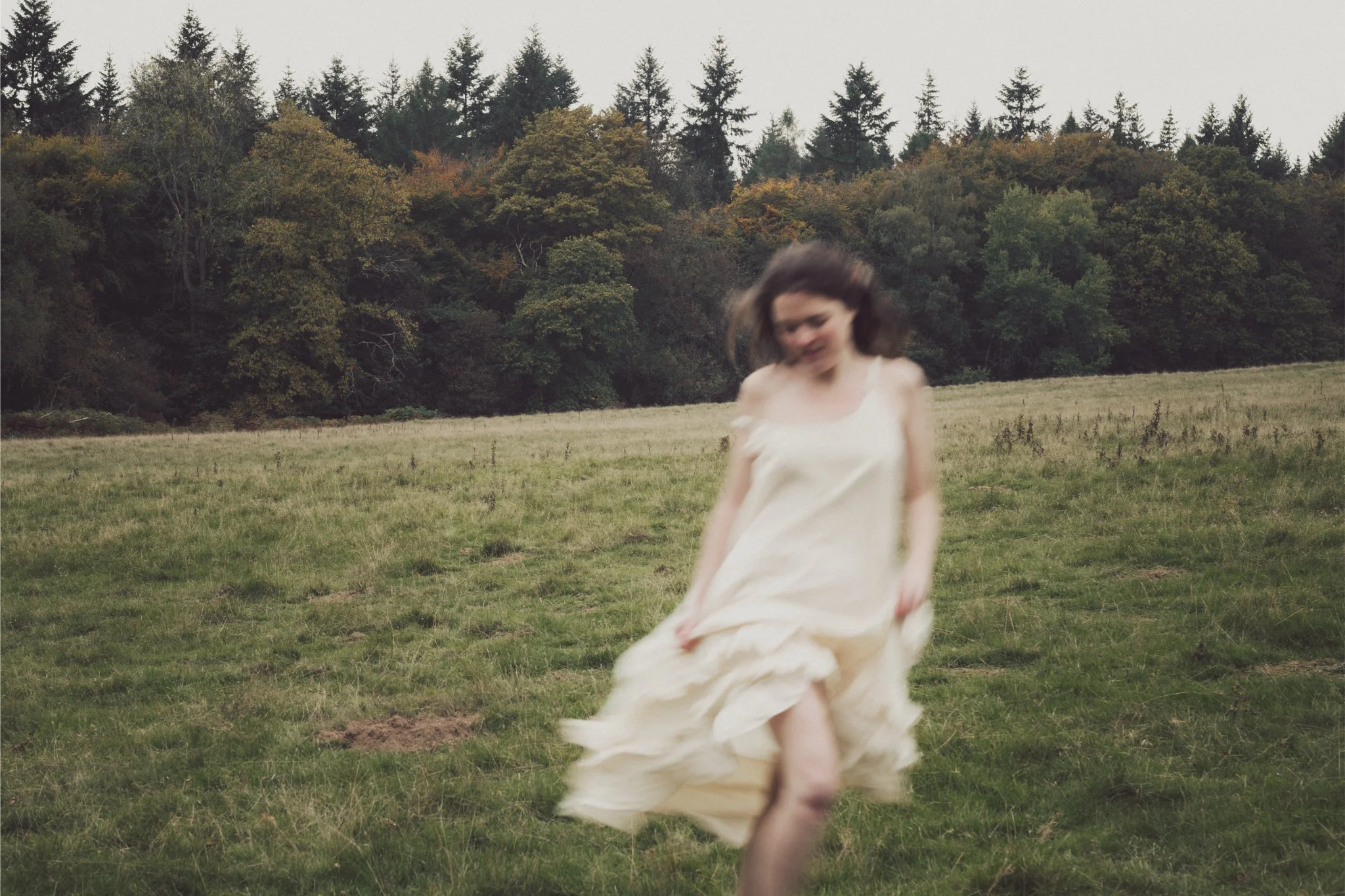 A woman wearing a light-colored dress is walking in a grassy field, with a backdrop of a forest of trees and a gray sky.