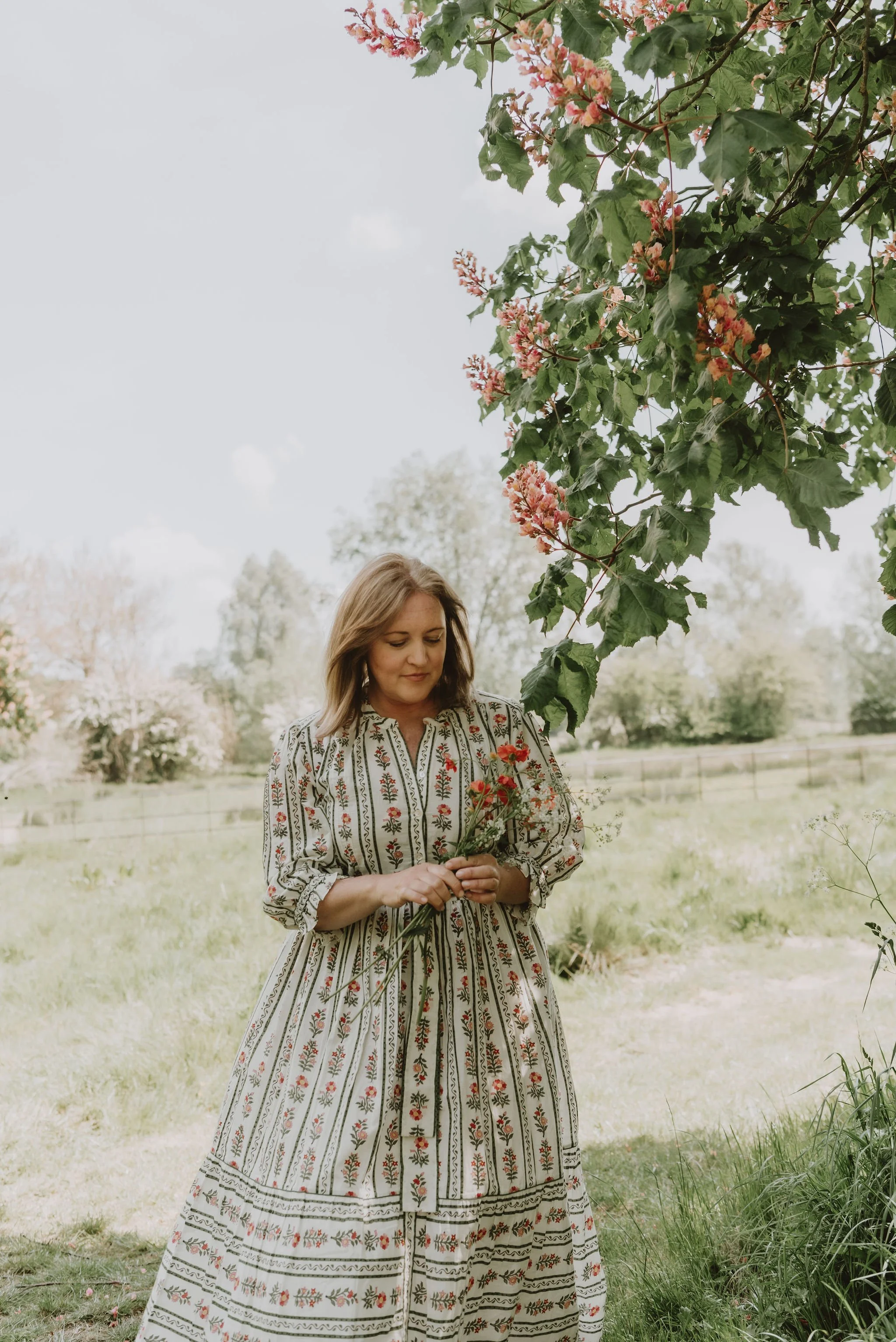 A woman in a patterned dress holding a small bouquet of red flowers in an outdoor setting with trees and a fence in the background.