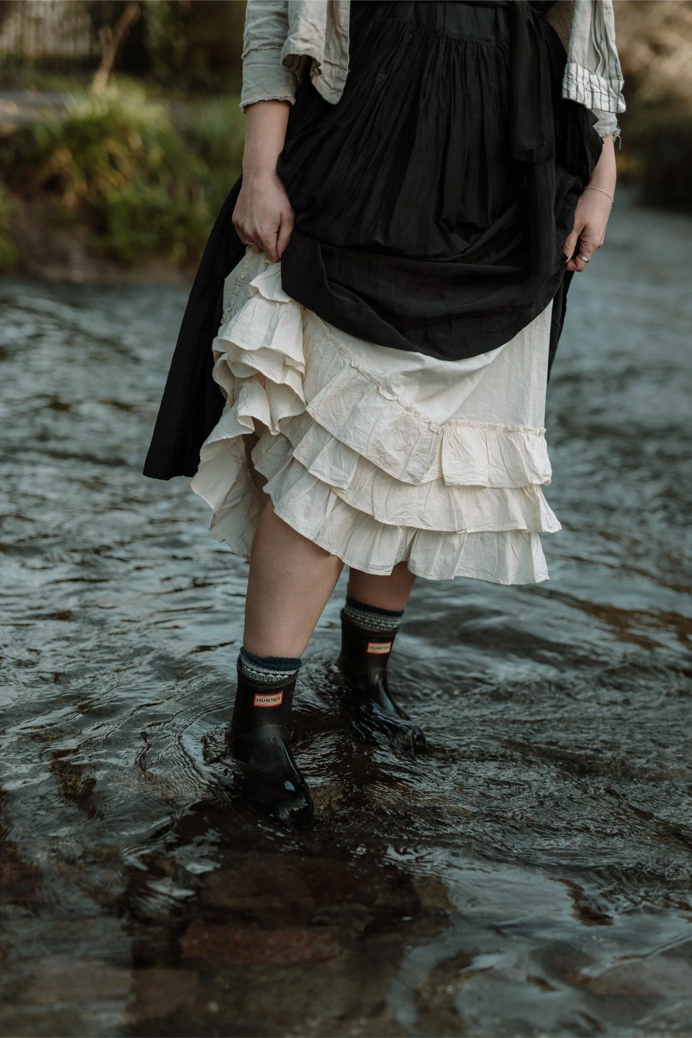 Person standing in a shallow stream, wearing a layered cream-colored skirt, black skirt, patterned socks, and waterproof boots.