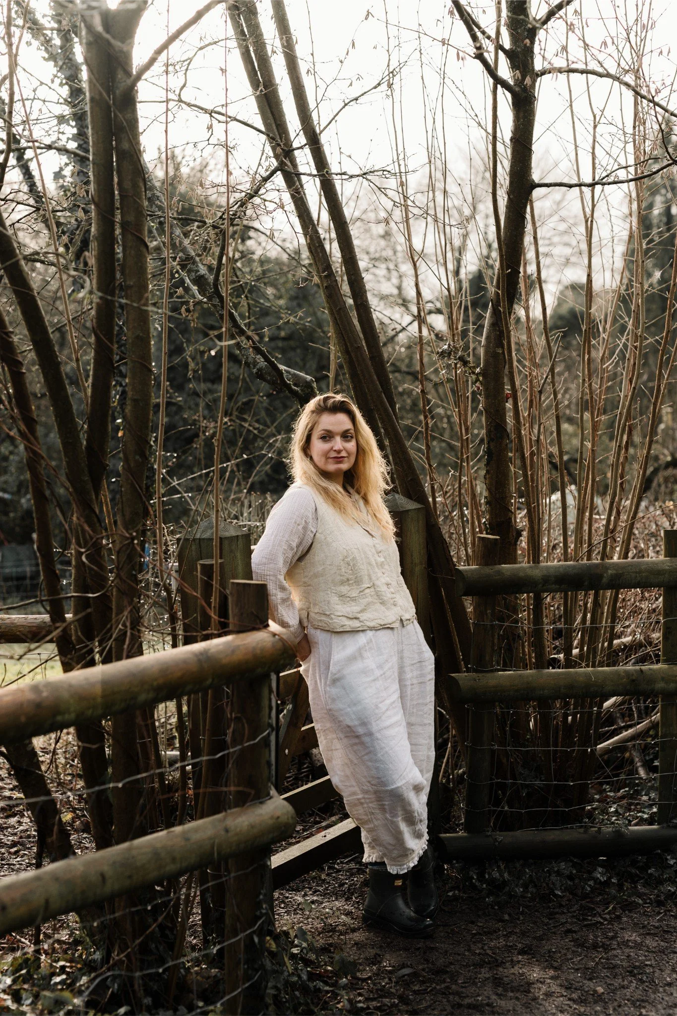 A woman with blonde hair, wearing a cream-colored long-sleeve top and loose cream-colored pants, stands outdoors on a dirt path next to a wooden fence, with leafless trees and bushes behind her.