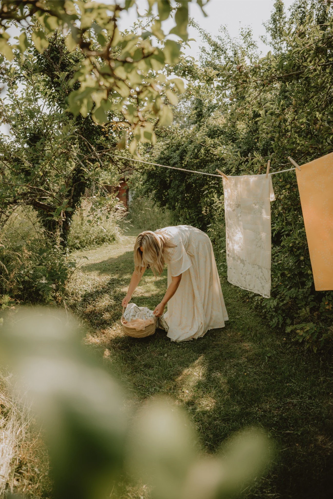 A woman in a light-colored dress bends down in a garden, reaching into a basket. There is a clothesline with fabric hanging on it, surrounded by greenery.