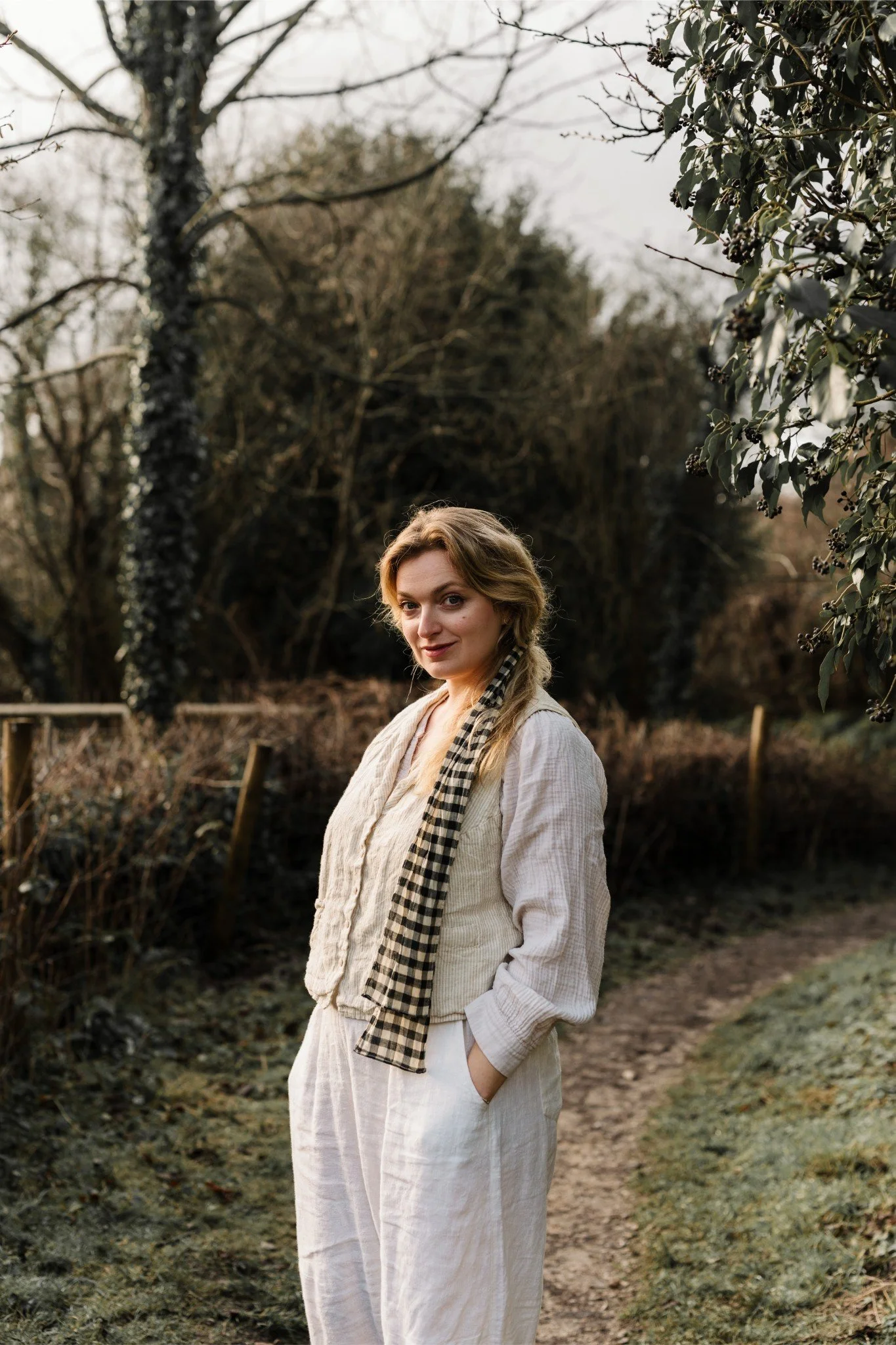 A woman with blonde hair in a braid, dressed in cream-colored clothing, stands outdoors on a dirt path surrounded by trees and bushes, with her hands in her pockets, looking at the camera.