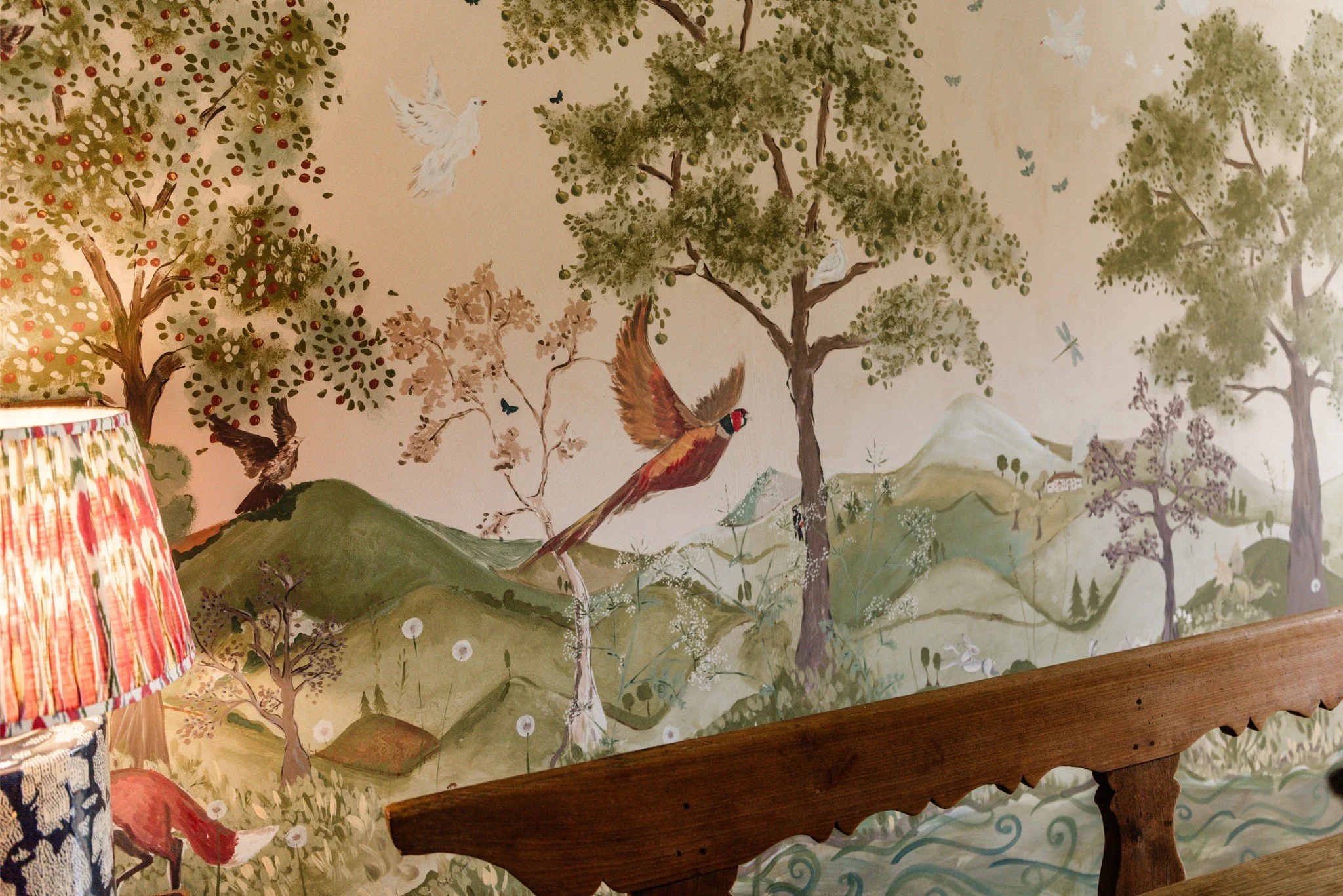 Painted mural of a nature scene with trees, mountains, birds, butterflies, and rolling hills in soft earthy tones, partially obscured by a wooden headboard and a colorful lampshade.