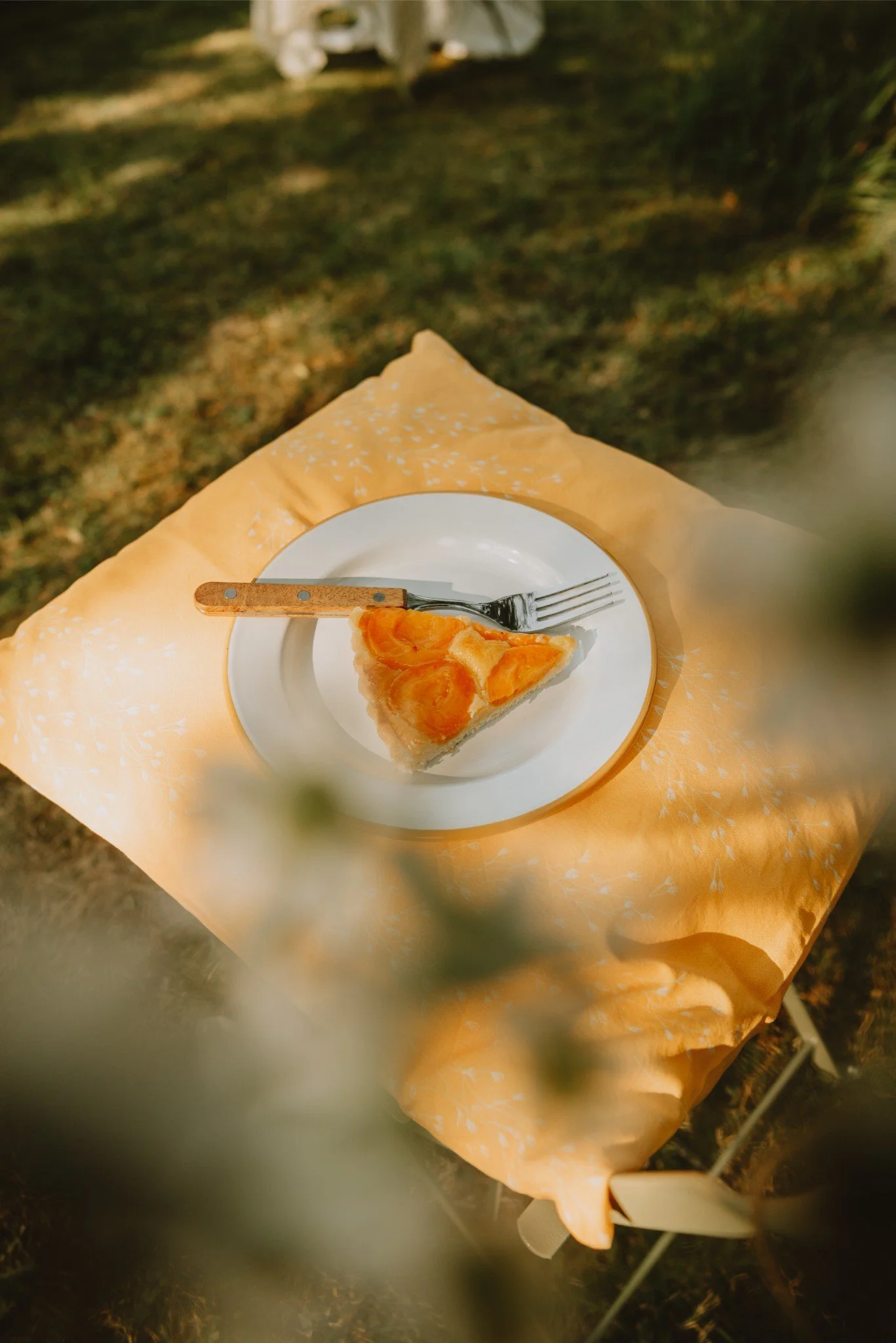 A slice of peach pie on a white plate with a fork and a small butter knife, placed on a yellow tablecloth outdoors.