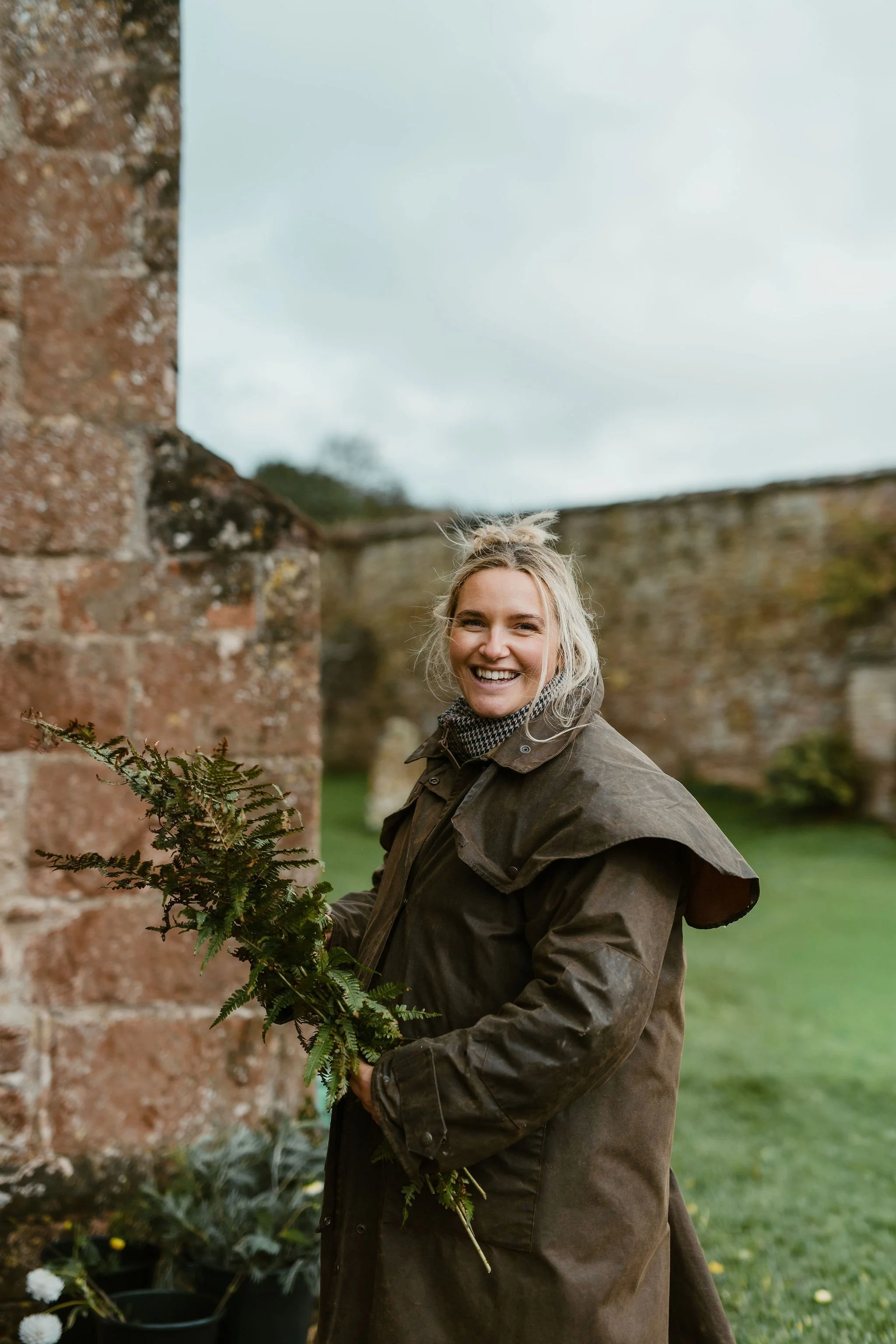 A woman with a big smile, wearing a brown raincoat, holding a green fern, standing outdoors near an old brick wall.