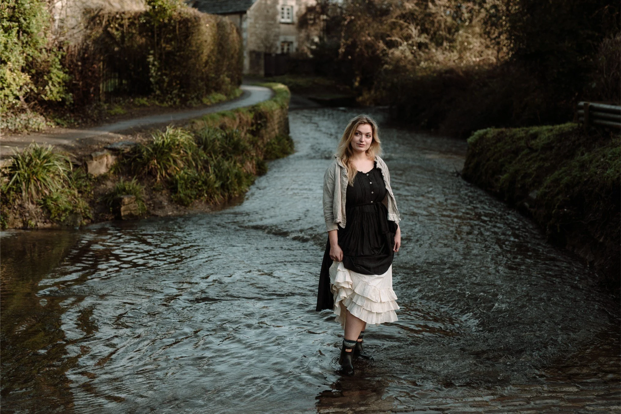A woman standing in a shallow stream with a rocky, mossy bank and a pathway in the background, surrounded by trees and bushes.