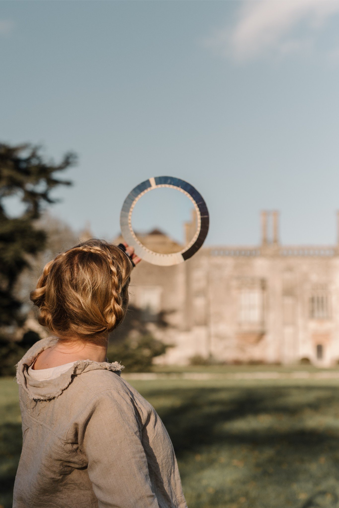 A woman with braided hair holding a reflective circular object outdoors in front of a historic building, with trees nearby and a clear sky above.