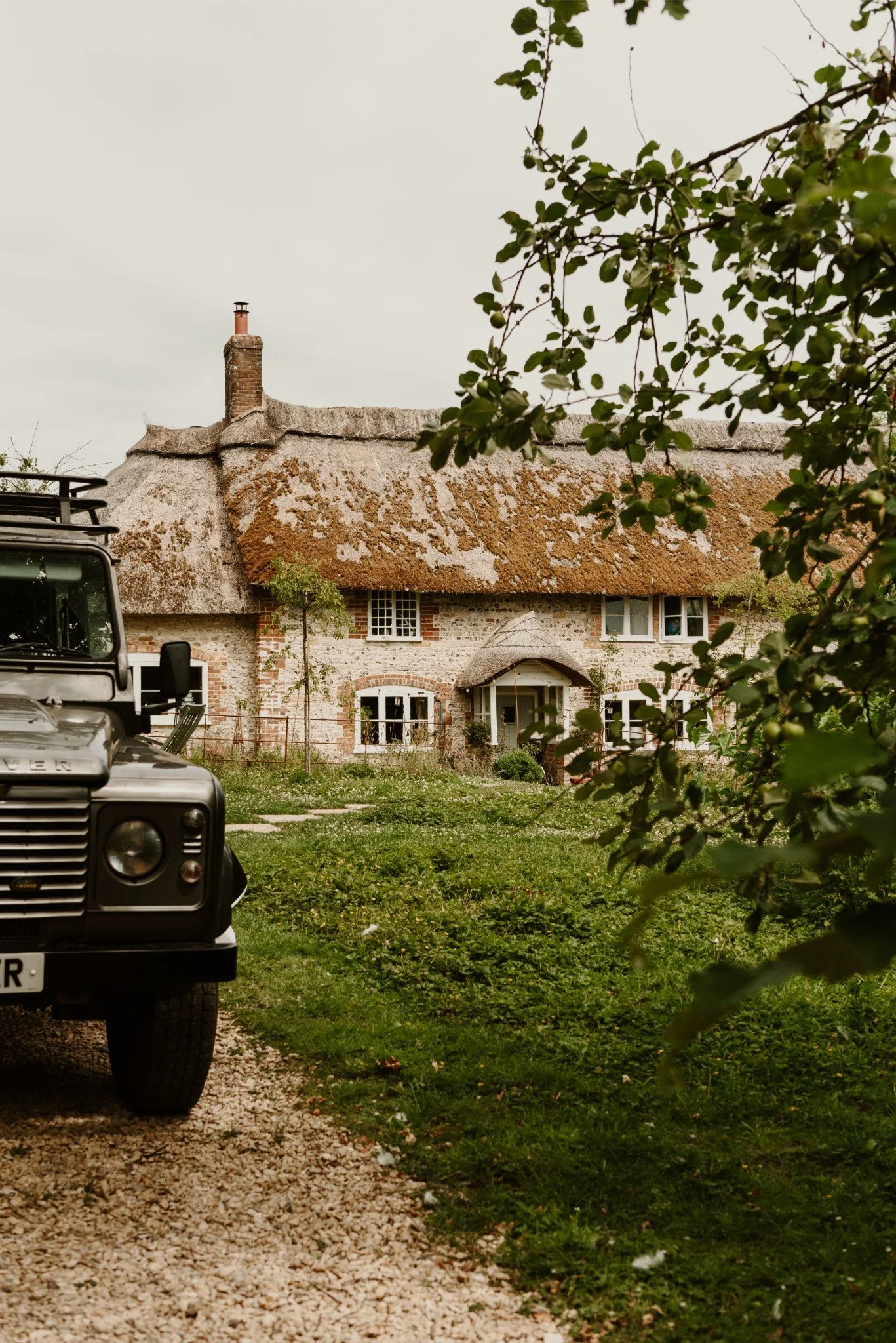 A rustic cottage with a thatched roof and brick walls, partially obscured by tree branches and leaves, with a vintage Land Rover parked on a gravel driveway in front.