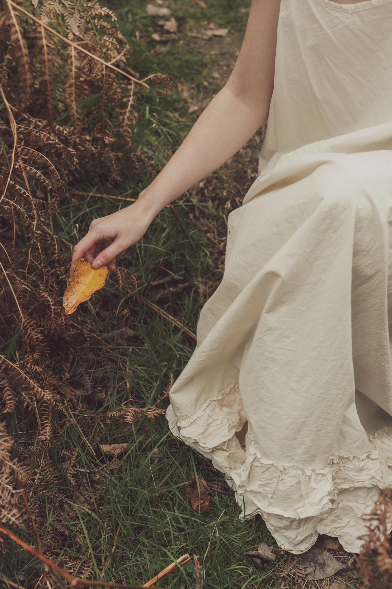 Person in a cream-colored dress bending down outdoors, holding a yellow autumn leaf among dry and green foliage.