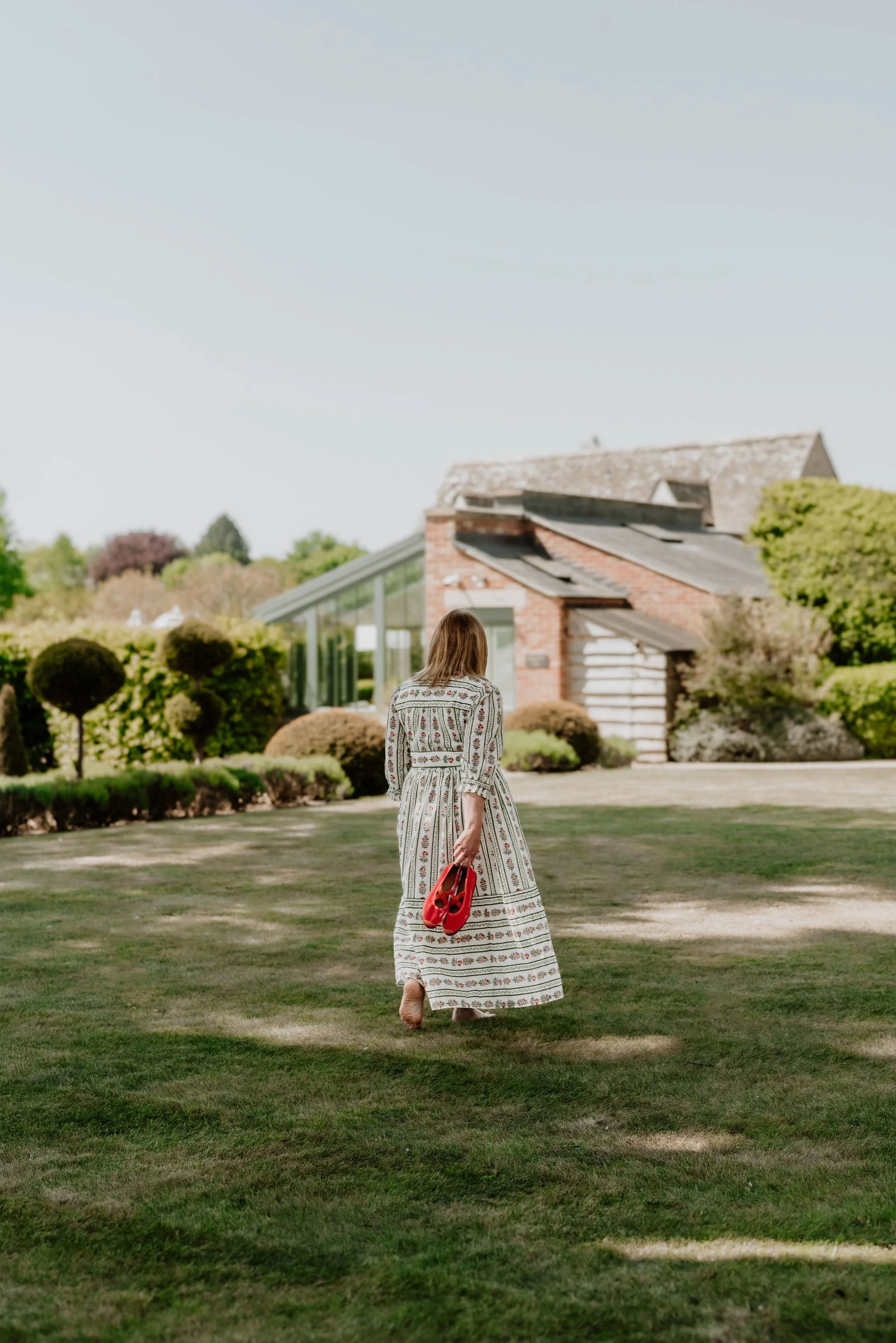 A woman walking on a lawn holding red shoes, with a house and greenery in the background.