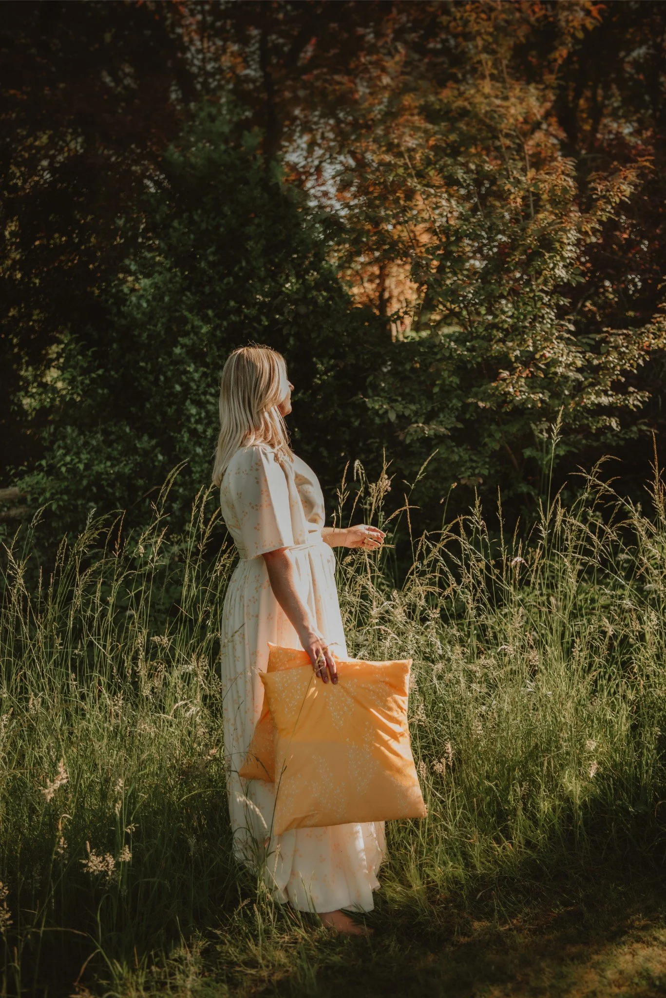 A woman in a light-colored dress standing in a grassy field with trees in the background, holding a large orange pillow.