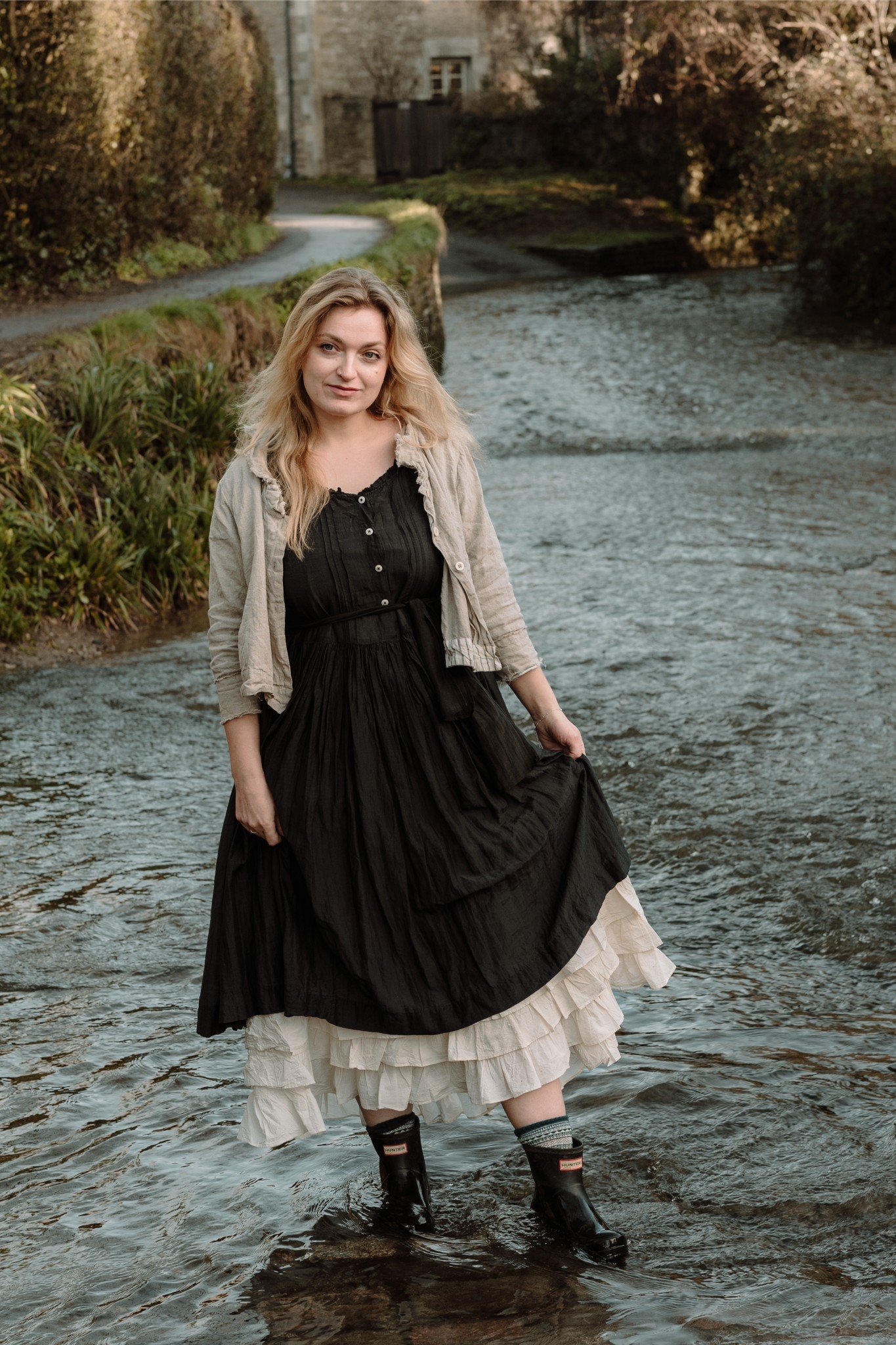 Woman standing in a stream, wearing a black dress, cream-colored petticoat, gray jacket, and rain boots, with trees and a house in the background.