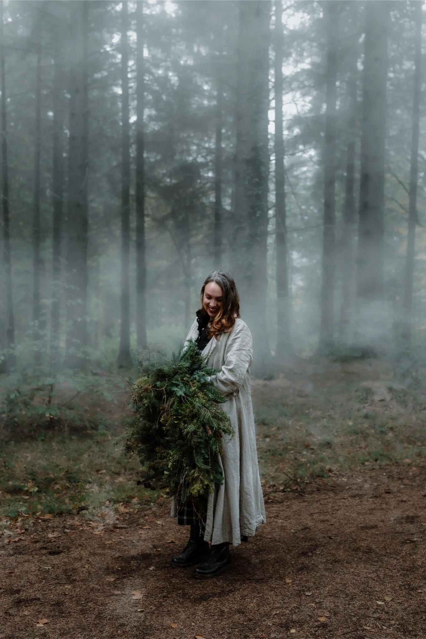 A woman standing in a misty forest, holding a large arrangement of pine branches and greenery, wearing a long beige coat and black boots, smiling and looking down.