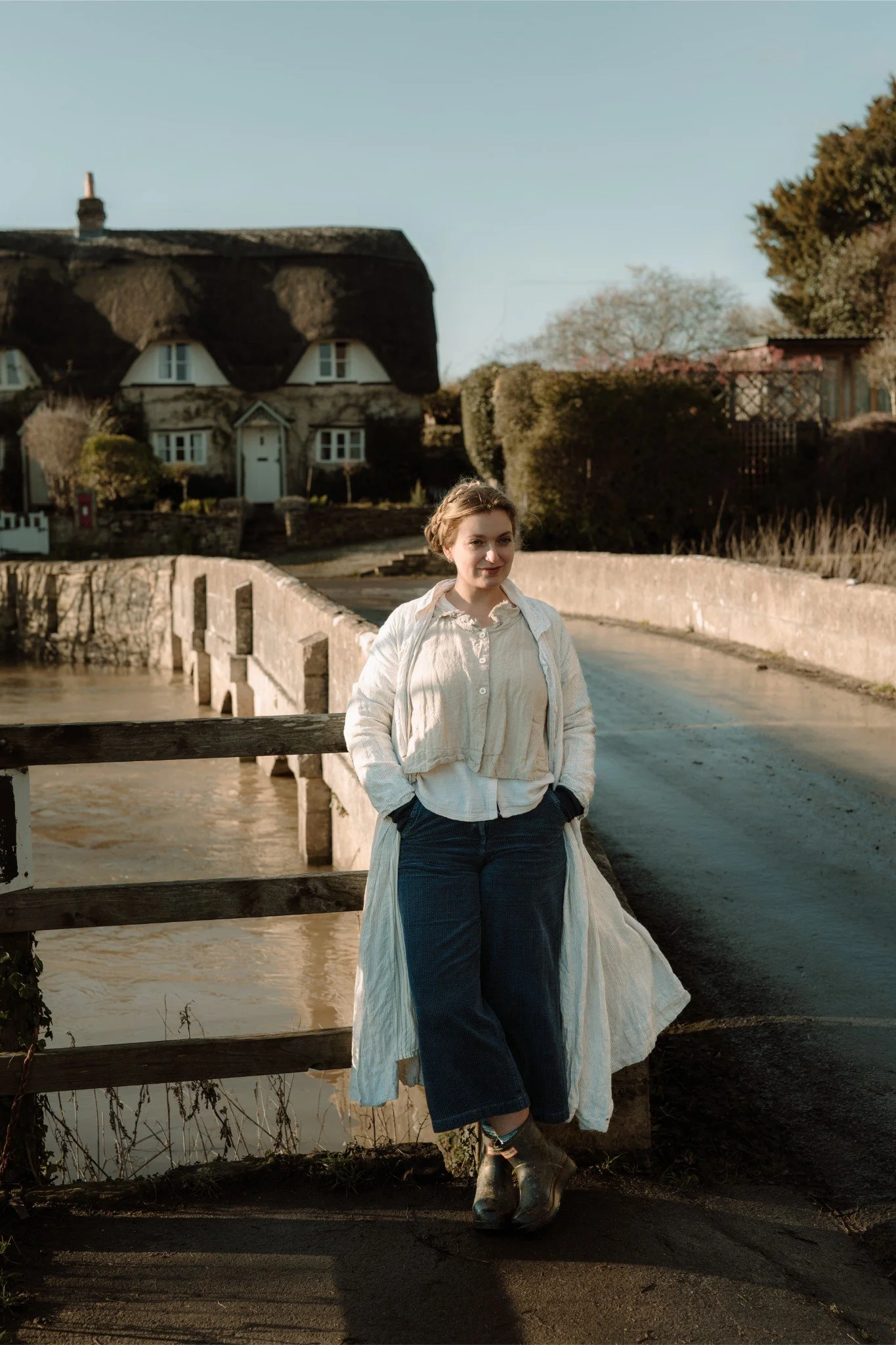A woman standing with hands in pockets on a small bridge over a river, with a thatched-roof cottage and trees in the background.
