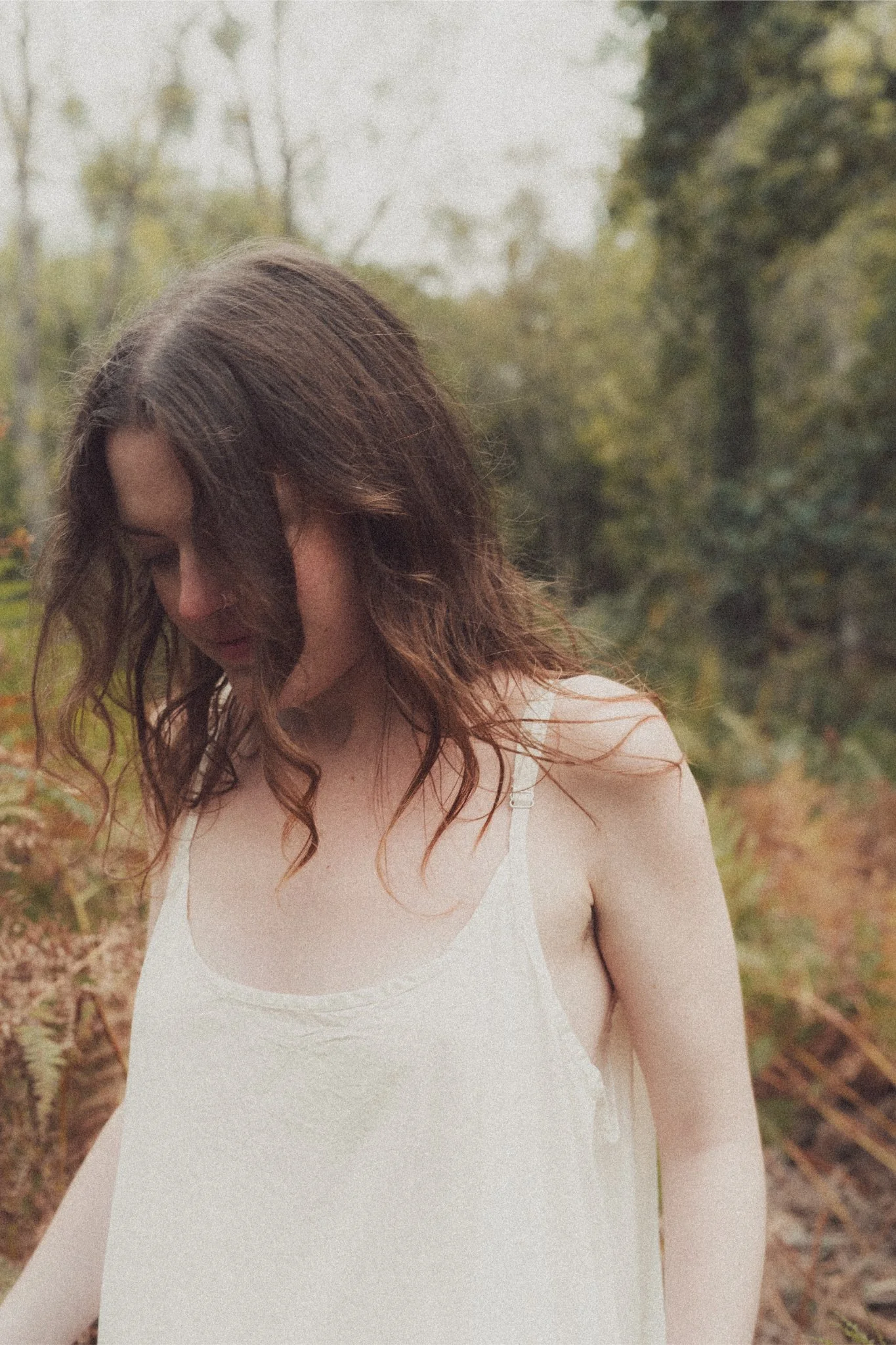 A woman with wavy brunette hair looking down, wearing a white slip dress, outdoors in a natural setting.