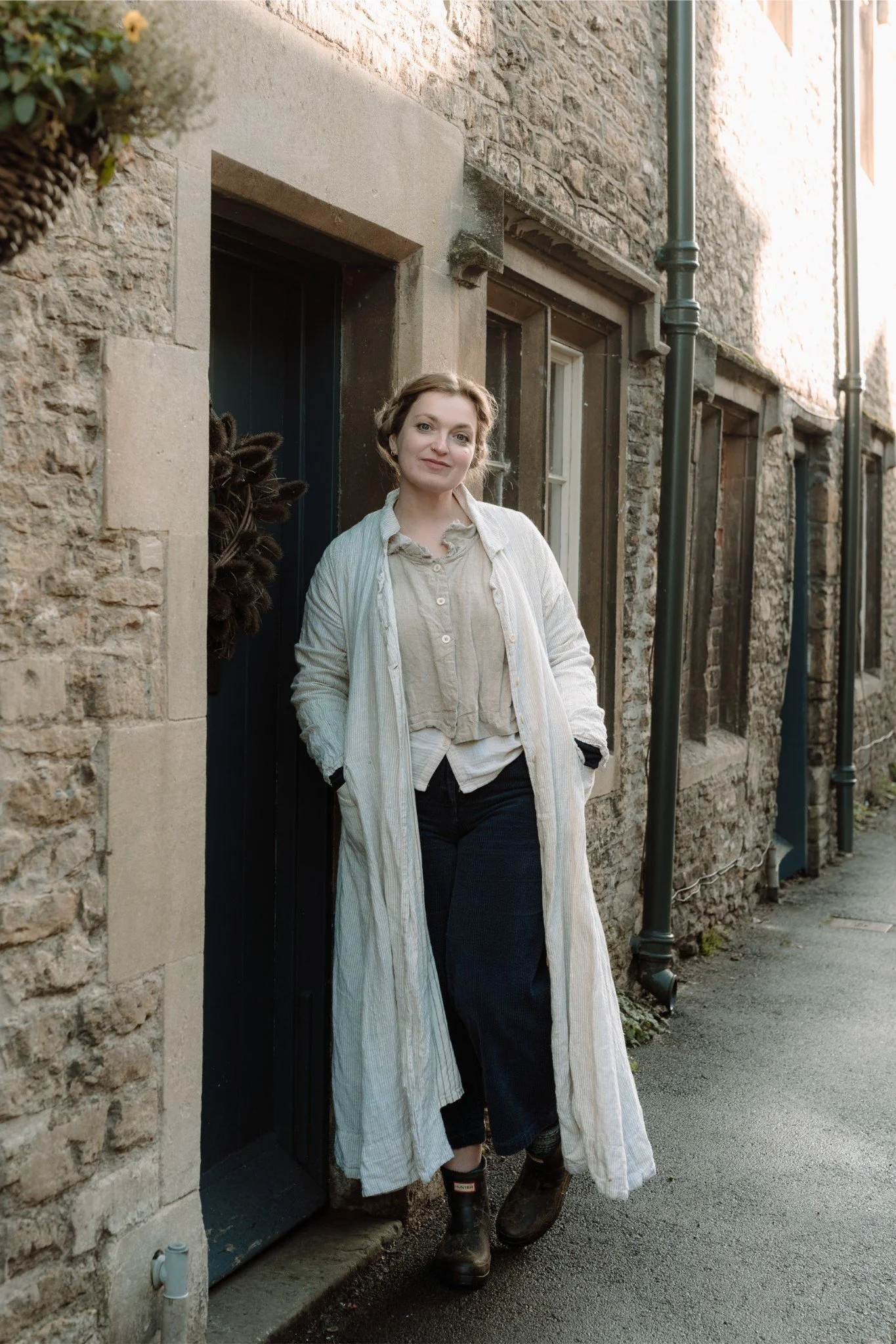 A woman standing outside a stone building with a black door, wearing a long striped coat, beige blouse, dark pants, and boots.
