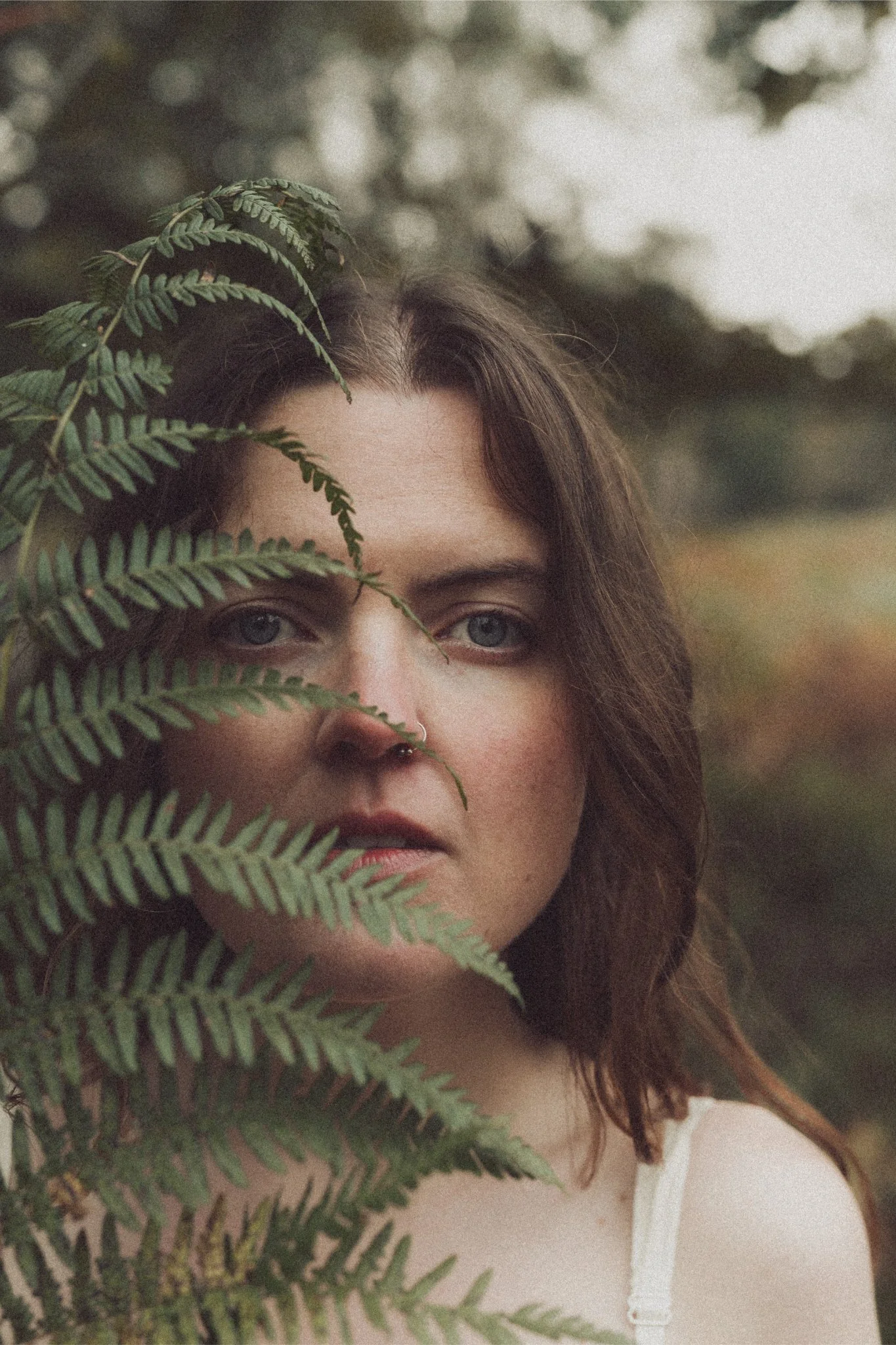 A woman with shoulder-length brown hair and fair skin is partially obscured by green fern leaves in an outdoor setting, with a blurred background of trees and sky.