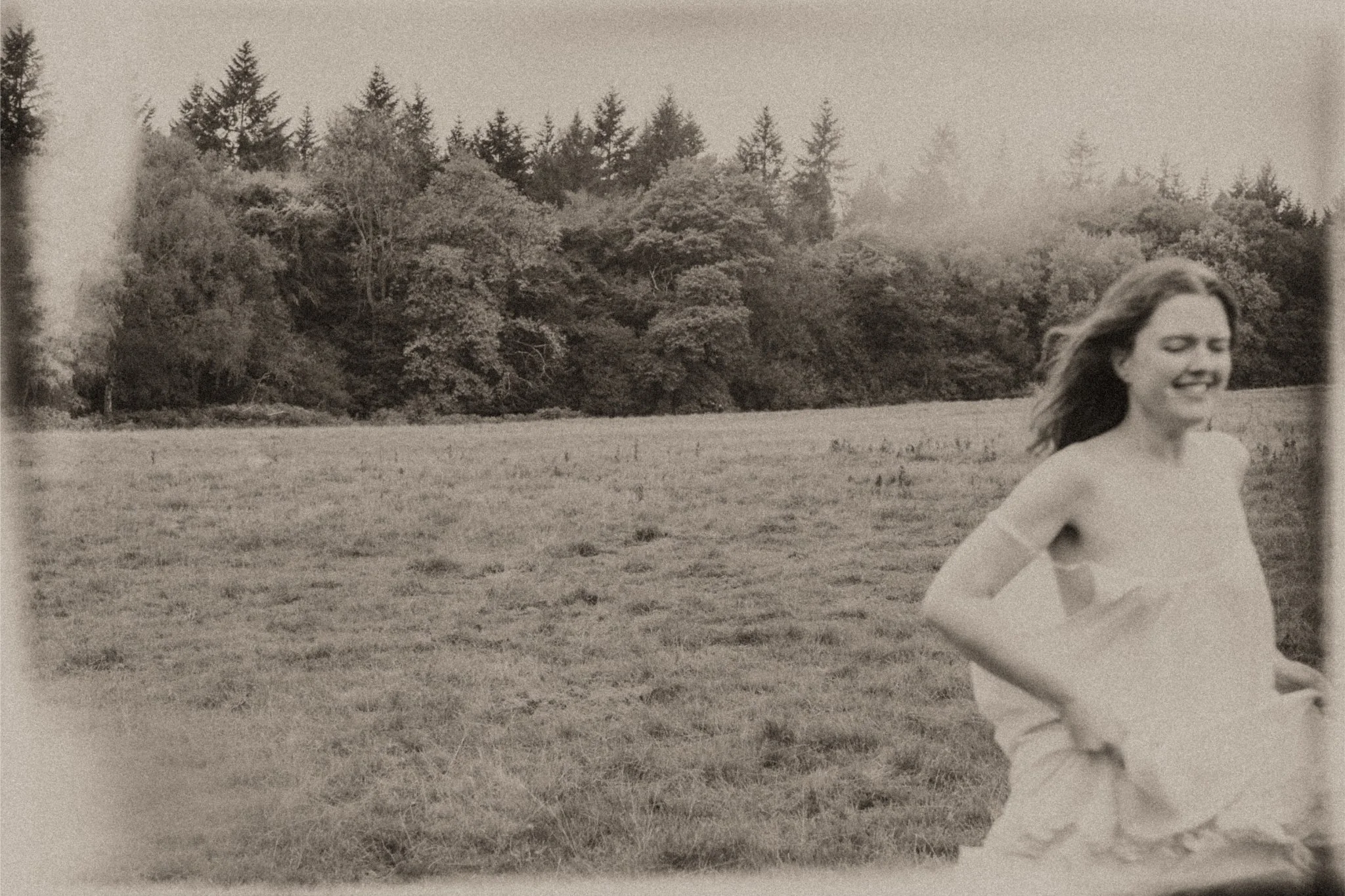 A woman with long hair running across a grassy field with a dense forest in the background, smiling and looking joyful.
