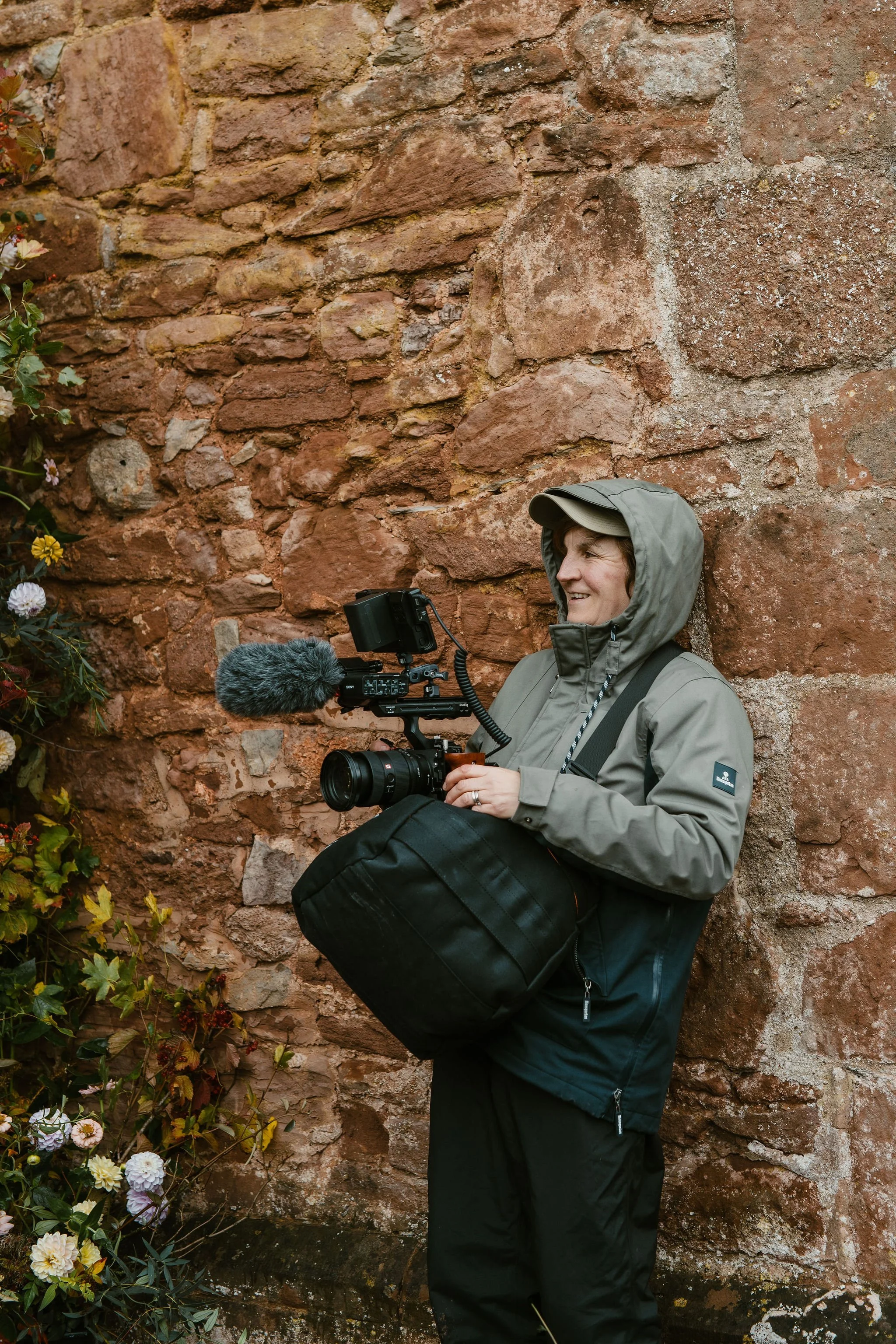 A person filming outdoors with a camera, microphone, and gear, standing against a weathered brick wall near flowers.