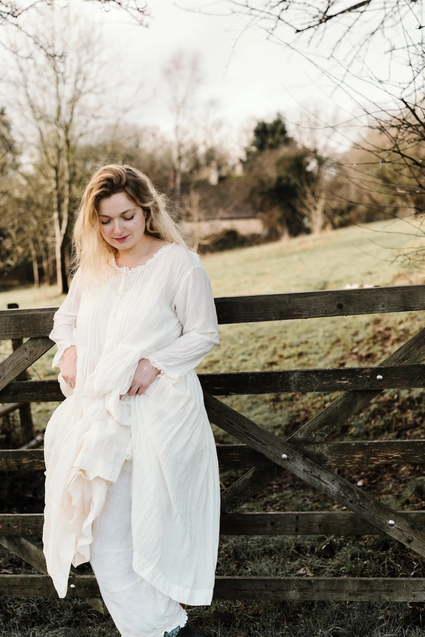 A young woman in a white dress standing by a wooden fence in an outdoor rustic setting during daytime.