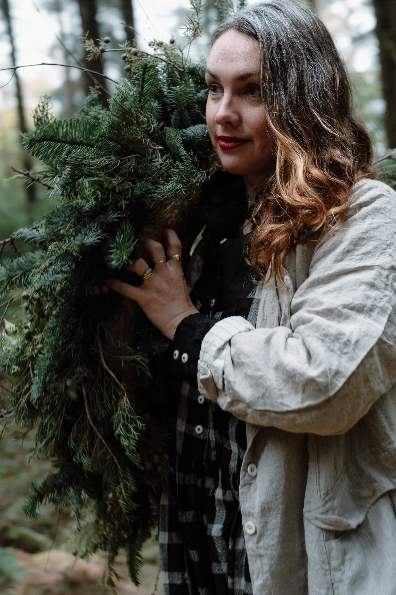 A woman in a forest holding a bundle of evergreen branches close to her face.