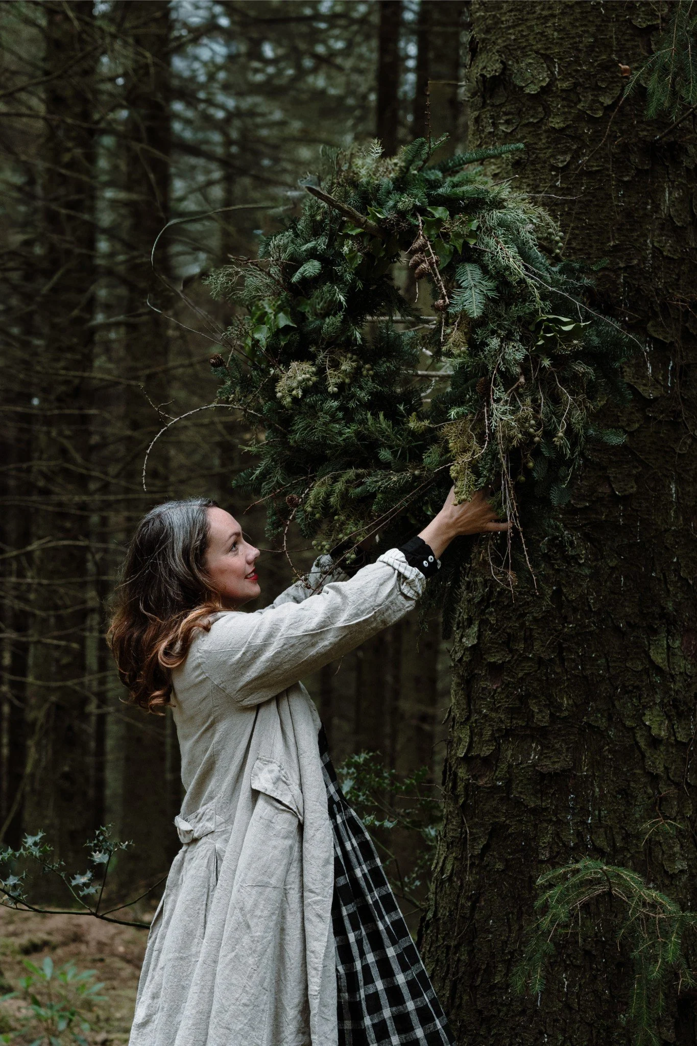 A woman placing a wreath made of pine branches on a tree in a forest.