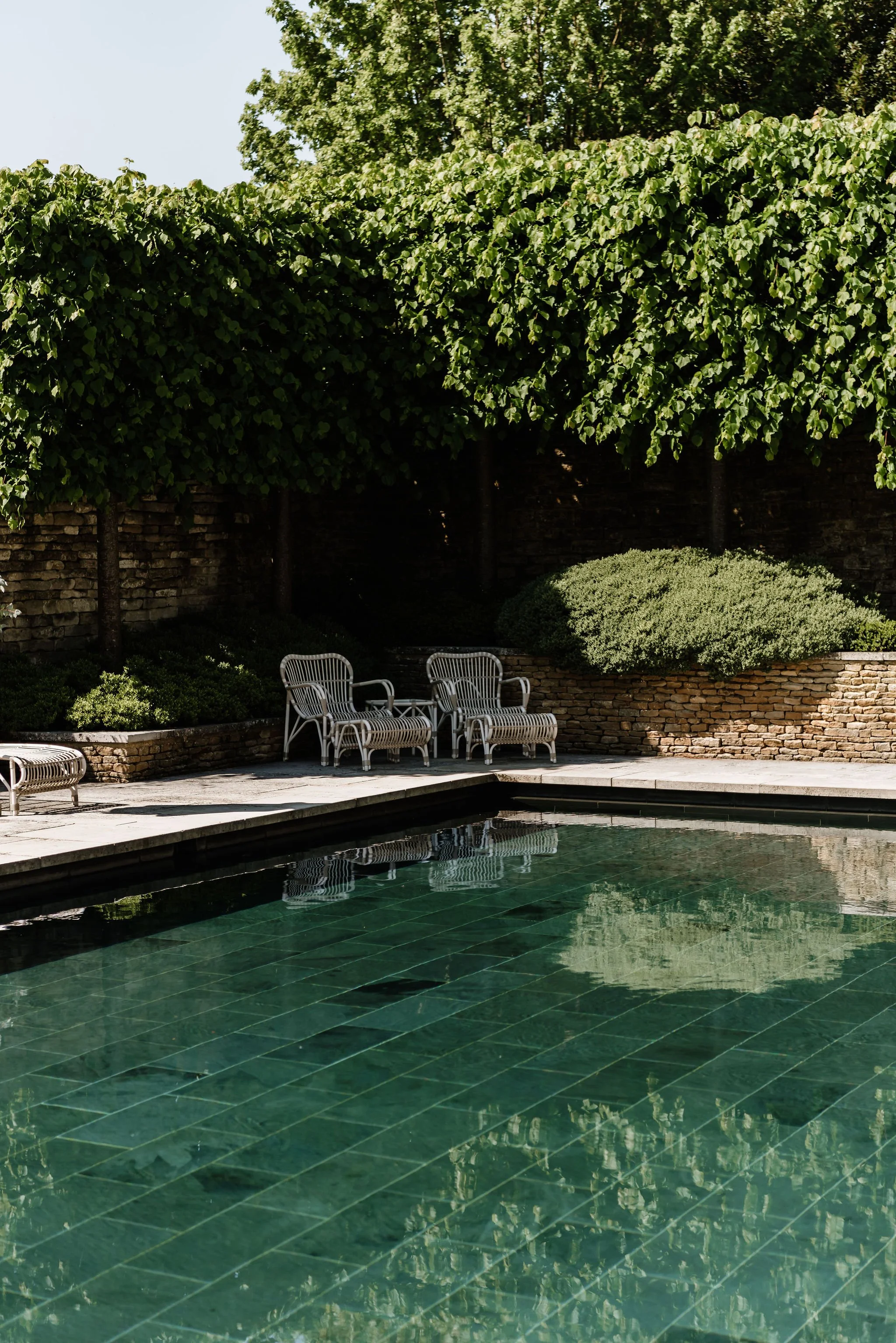 Poolside seating area with metal chairs and a bench, surrounded by greenery and a stone wall.