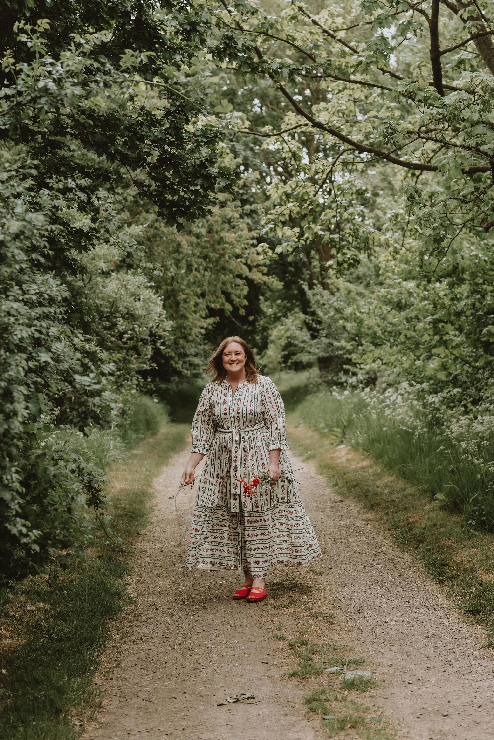 A woman in a patterned dress and red shoes walking along a dirt path surrounded by green trees and foliage, smiling and holding a small bouquet of red flowers.