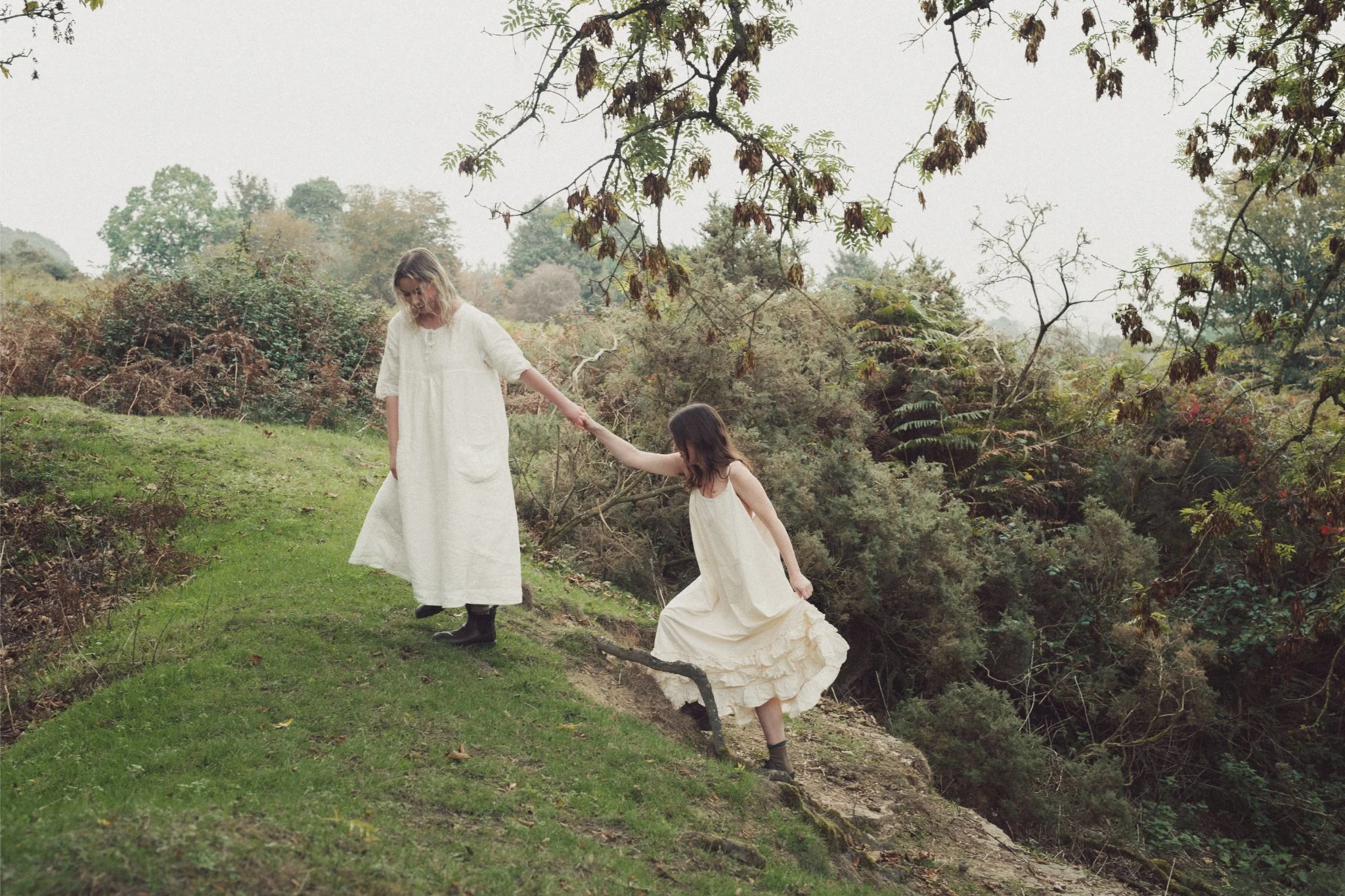 A woman and a girl, both in white dresses, walking along a grassy path on a hillside, holding hands and helping each other as they descend a slope in a lush, wooded area.